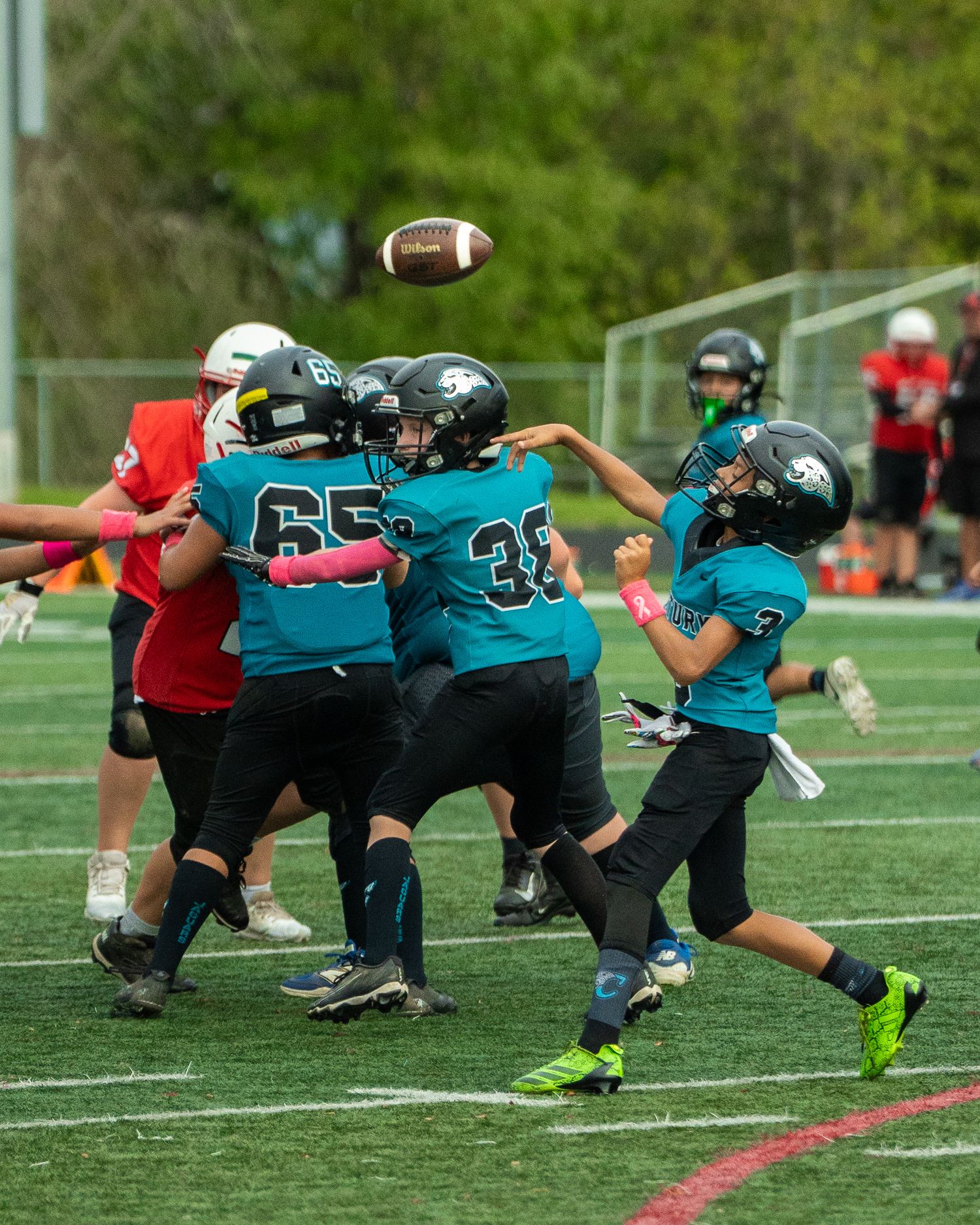 Youth football player in teal jersey performs stretching exercises on a grass field during practice.