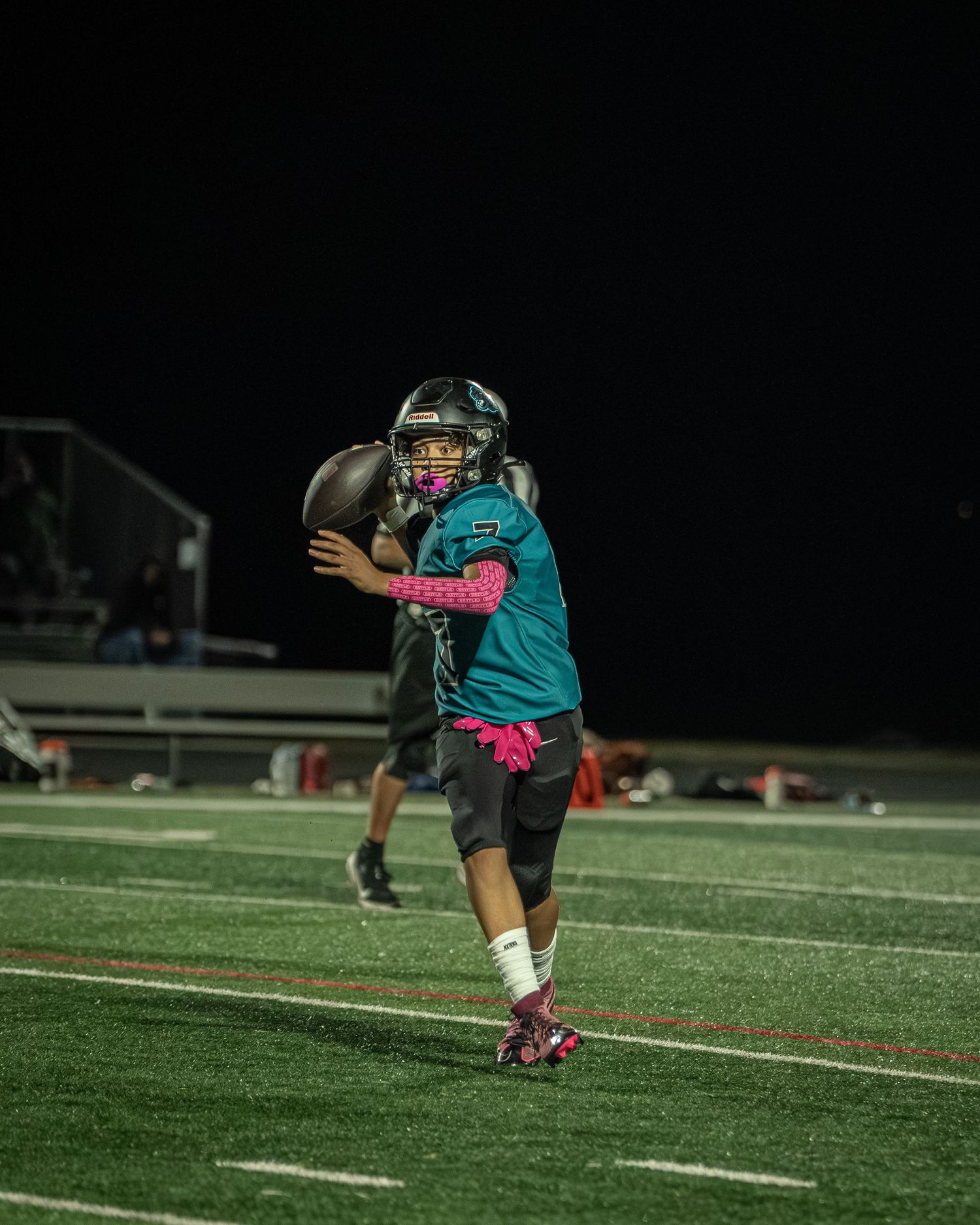 Players in teal jerseys perform dynamic stretching exercises on a football field at night.
