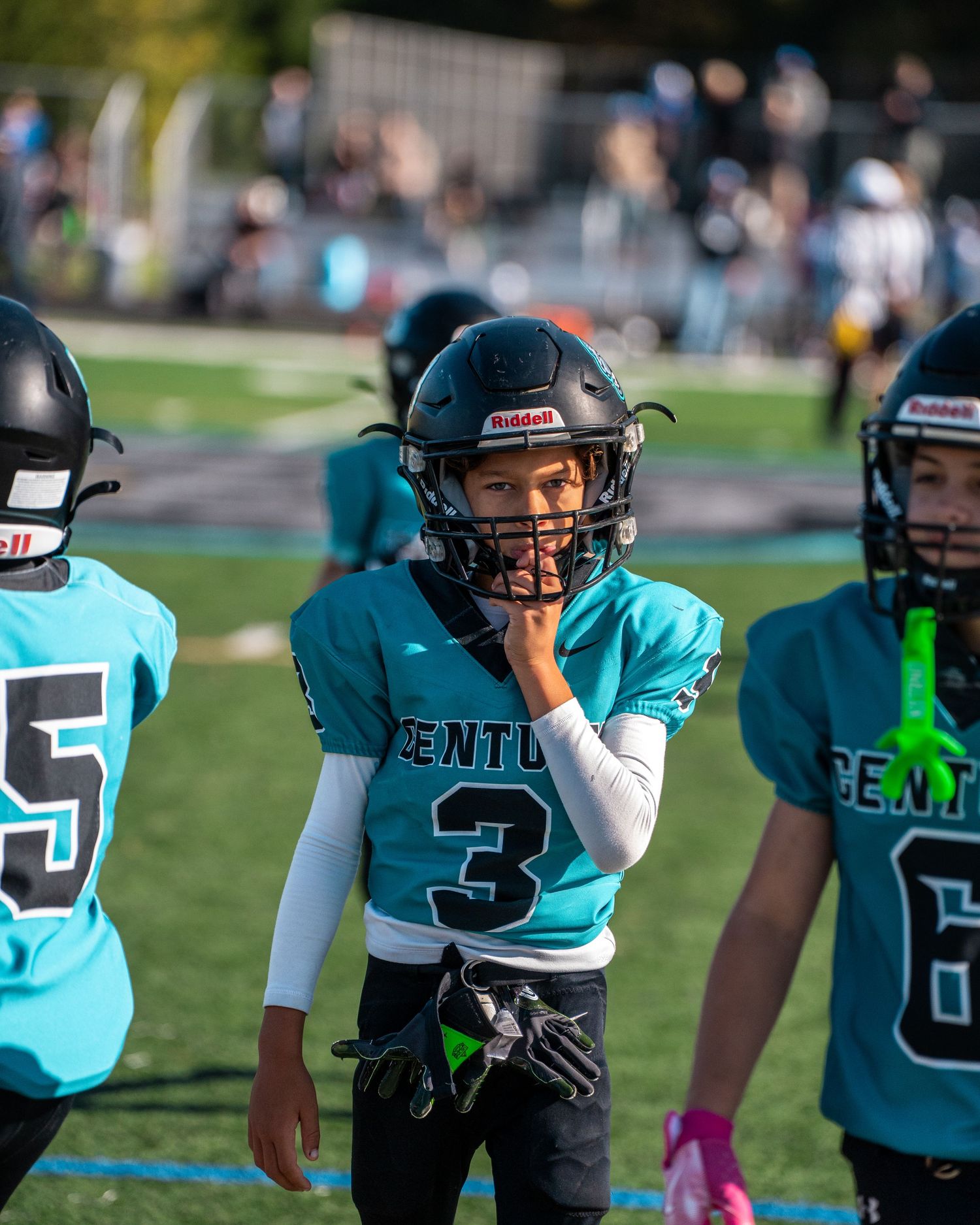 Youth football team in teal uniforms huddles on sideline during game.