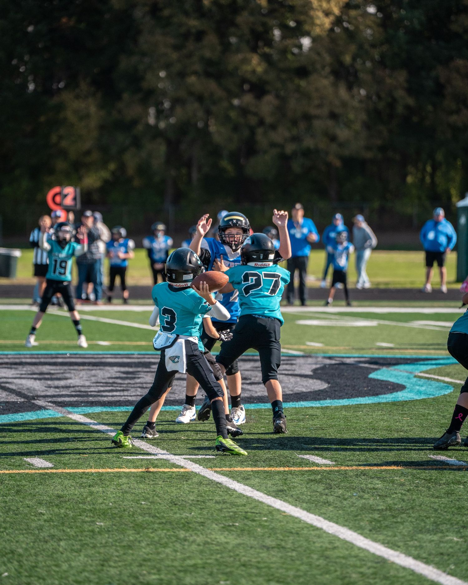 Football players in teal and black uniforms line up in formation on a sunny field during practice.