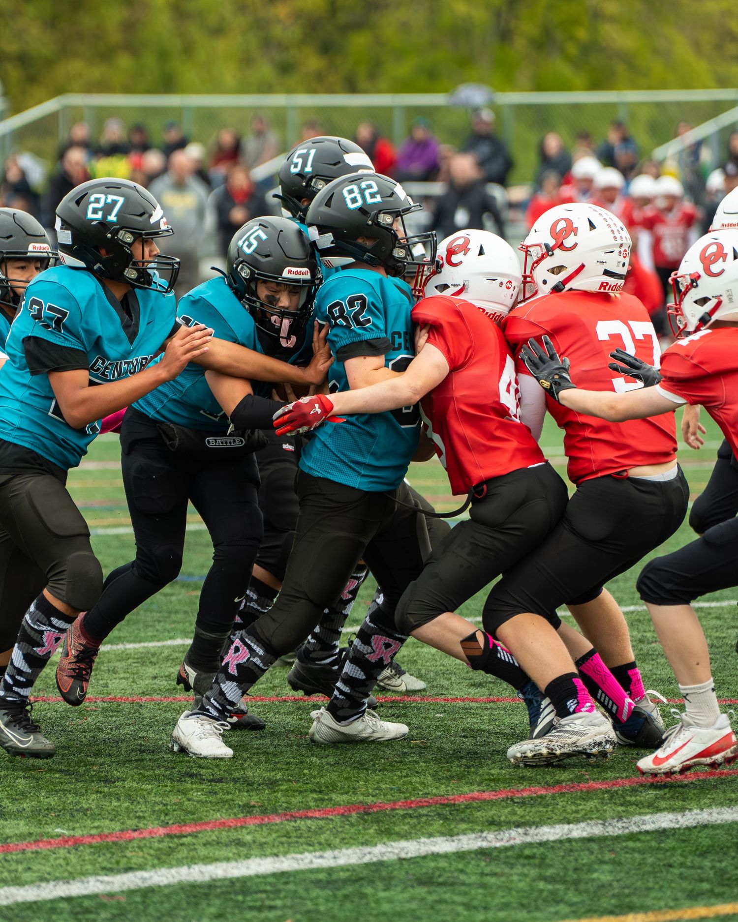 Two football teams line up at scrimmage with players in teal and red uniforms facing off.