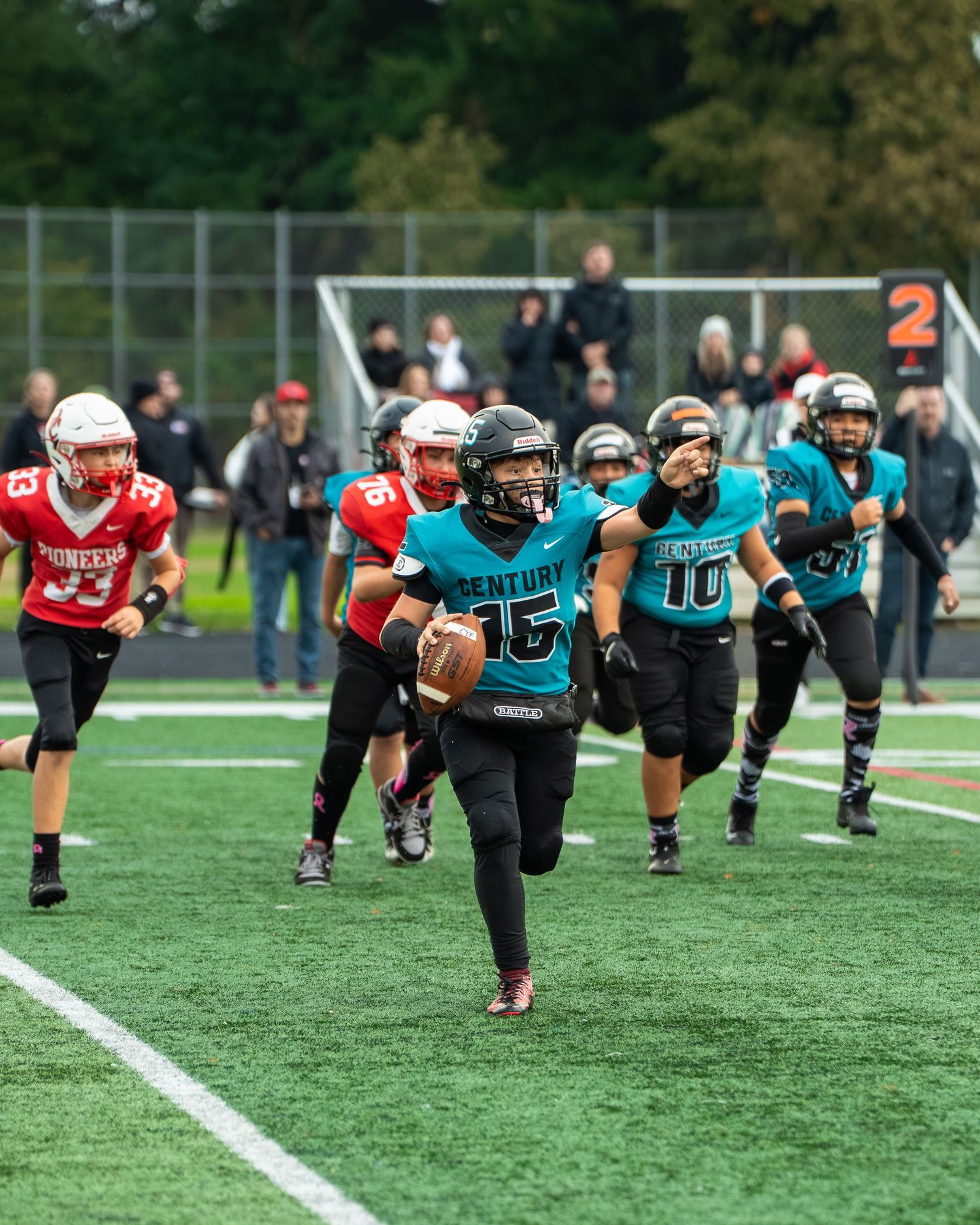 Football teams in teal and red uniforms compete during match on artificial turf field.