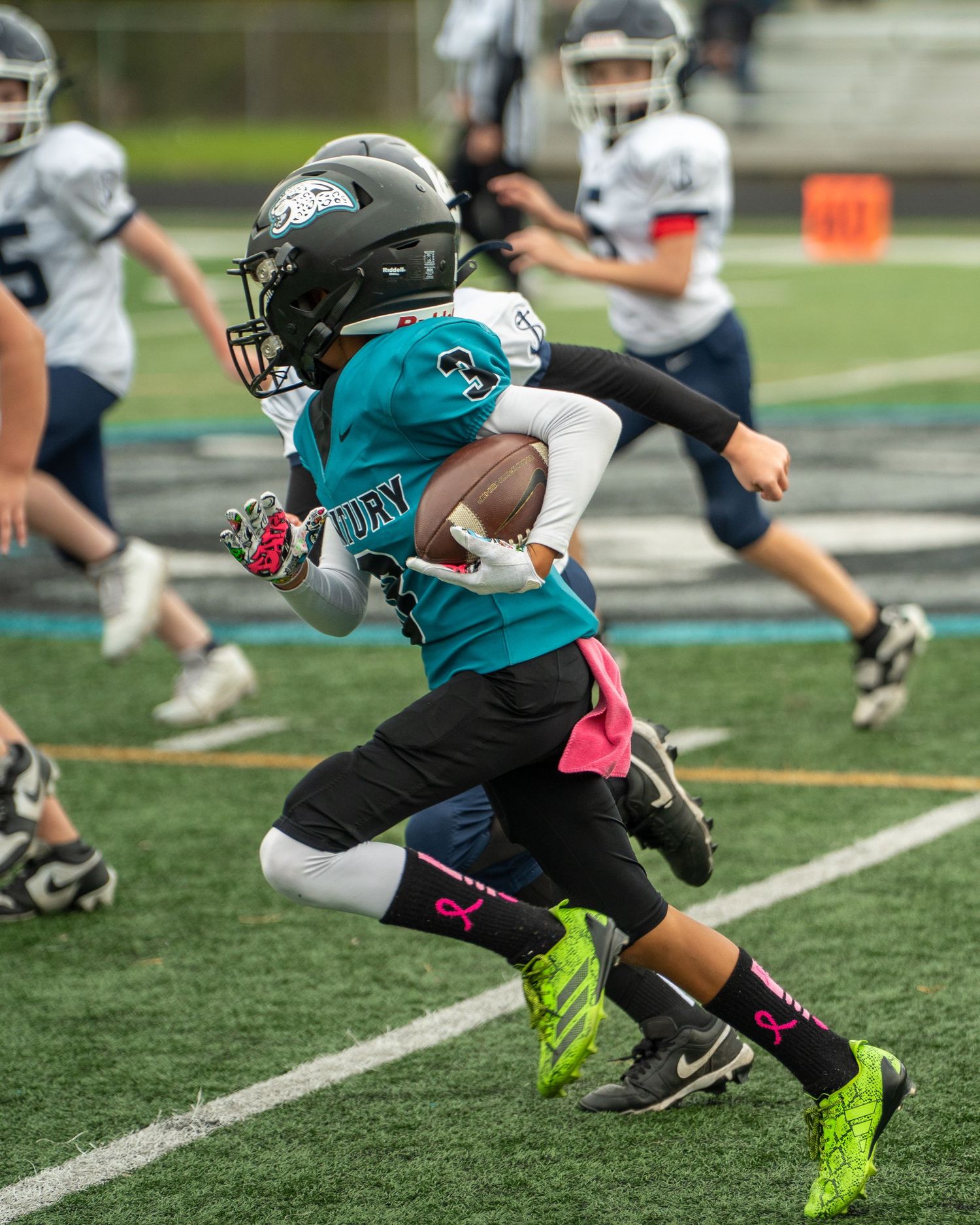 Youth athletes in teal and neon cleats sprint during football practice.
