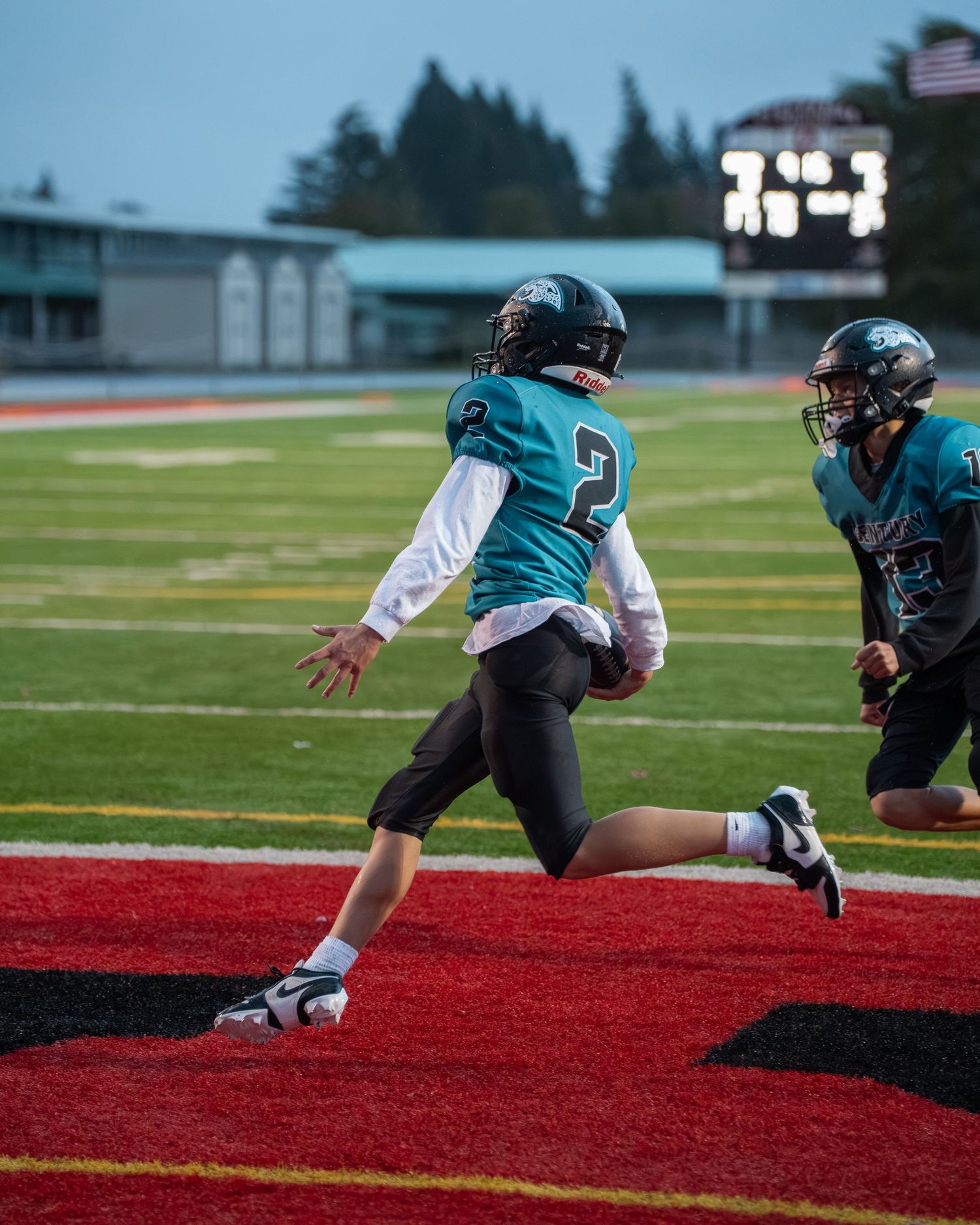 Football player in teal jersey diving for the end zone on red turf during a game.