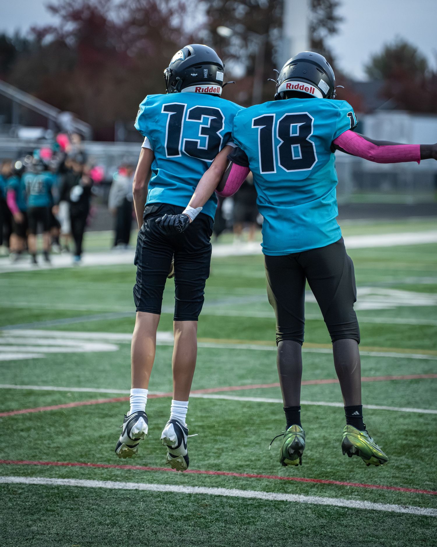 Football players in teal and black uniforms warm up along the sideline during practice.