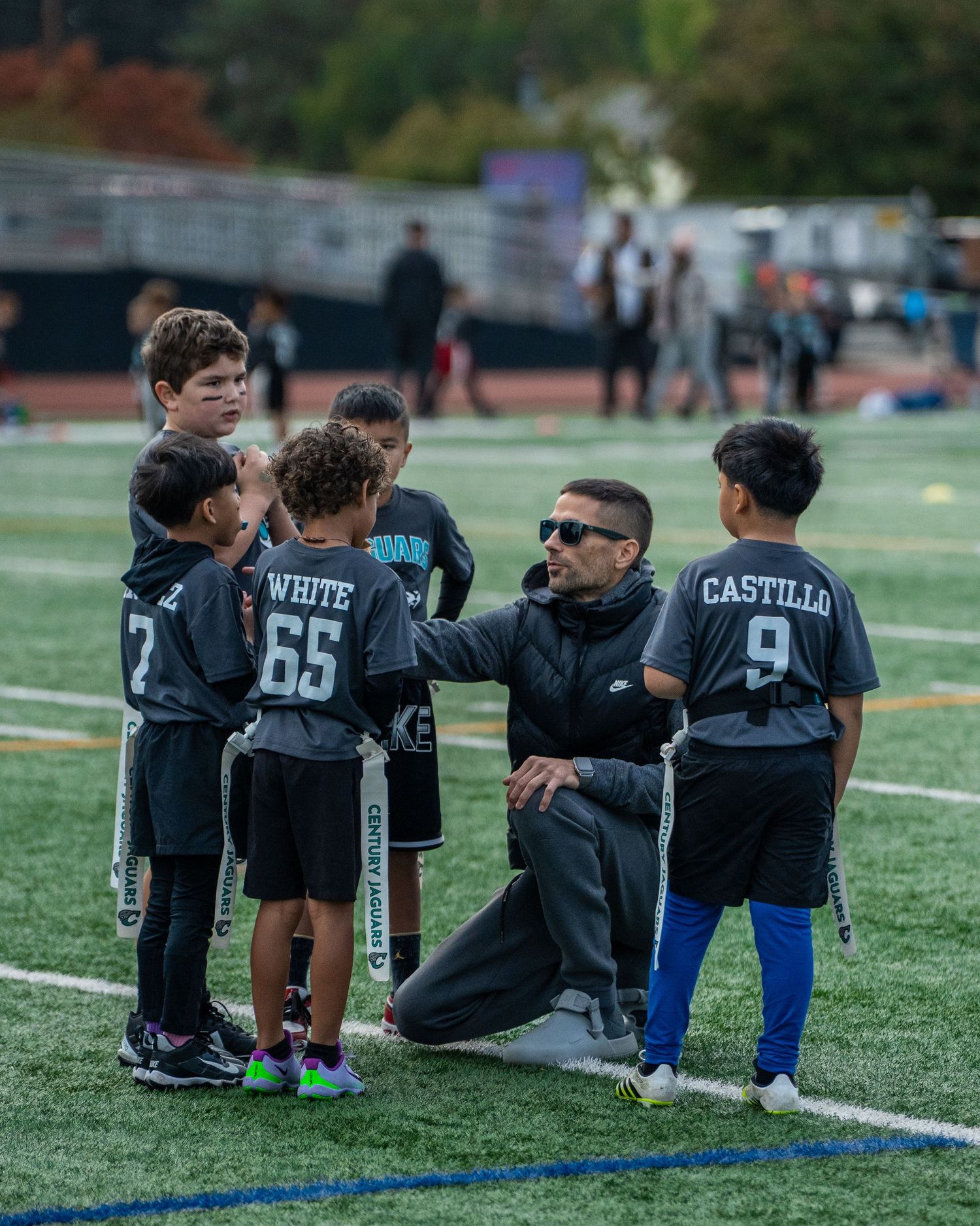 Youth football players in black jerseys sitting together on the sideline of a sports field.