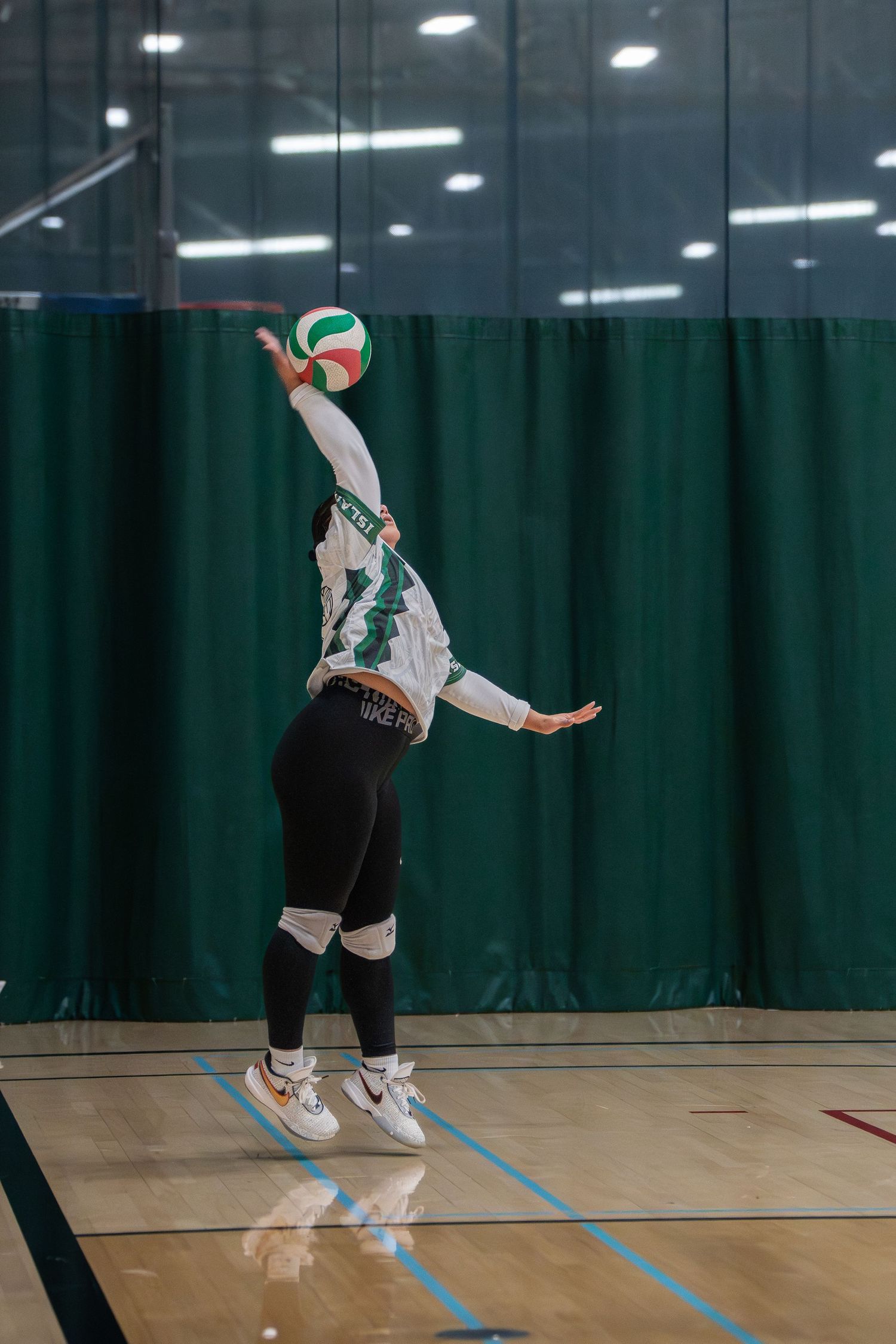 Player leaping to hit a volleyball in an indoor gymnasium with green curtains.