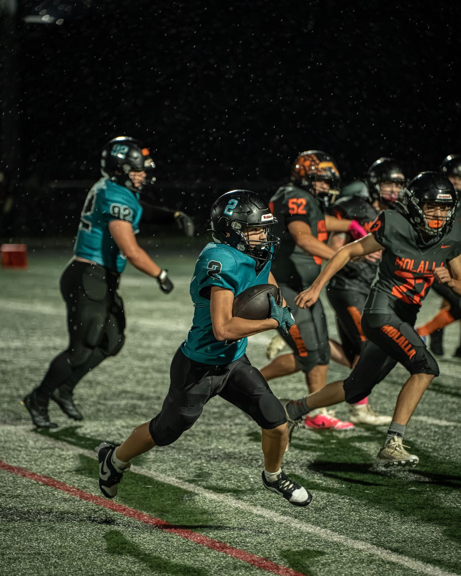 Running back in teal jersey carries ball through defensive line during night game.