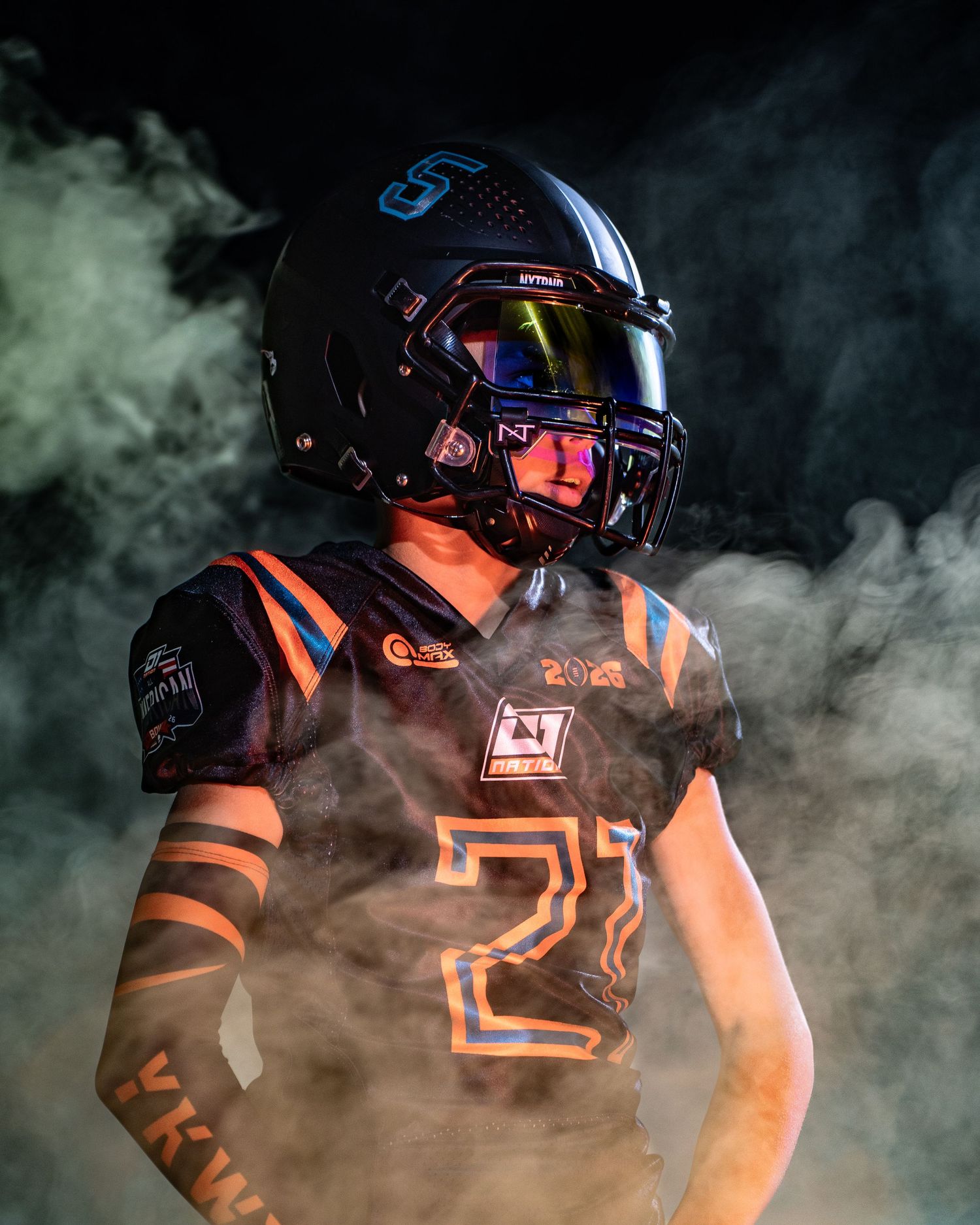 Football player in black uniform emerges through dramatic smoke effect with orange lighting.