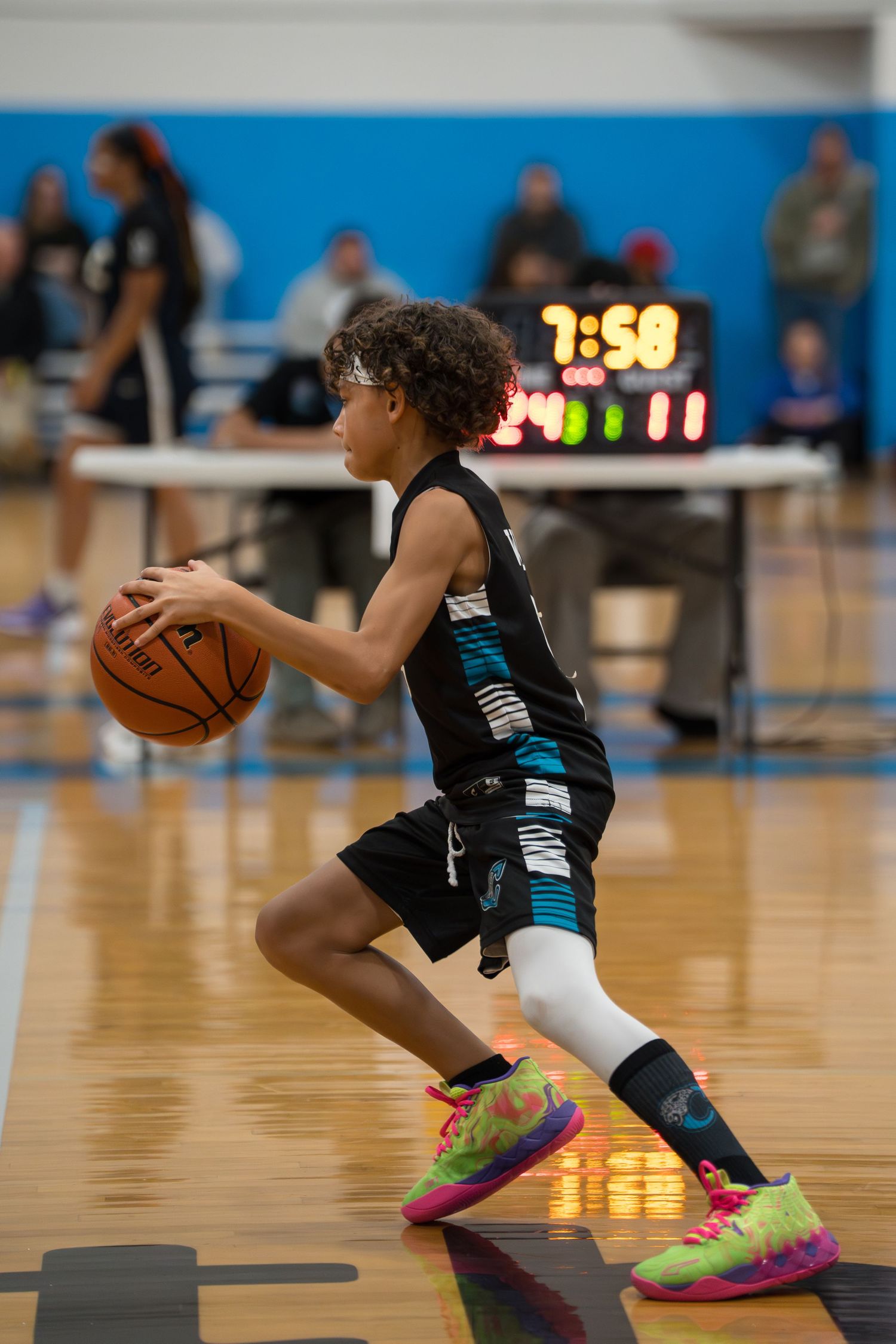 Basketball player in black uniform dribbles low while making quick moves on a hardwood court with neon lights visible.