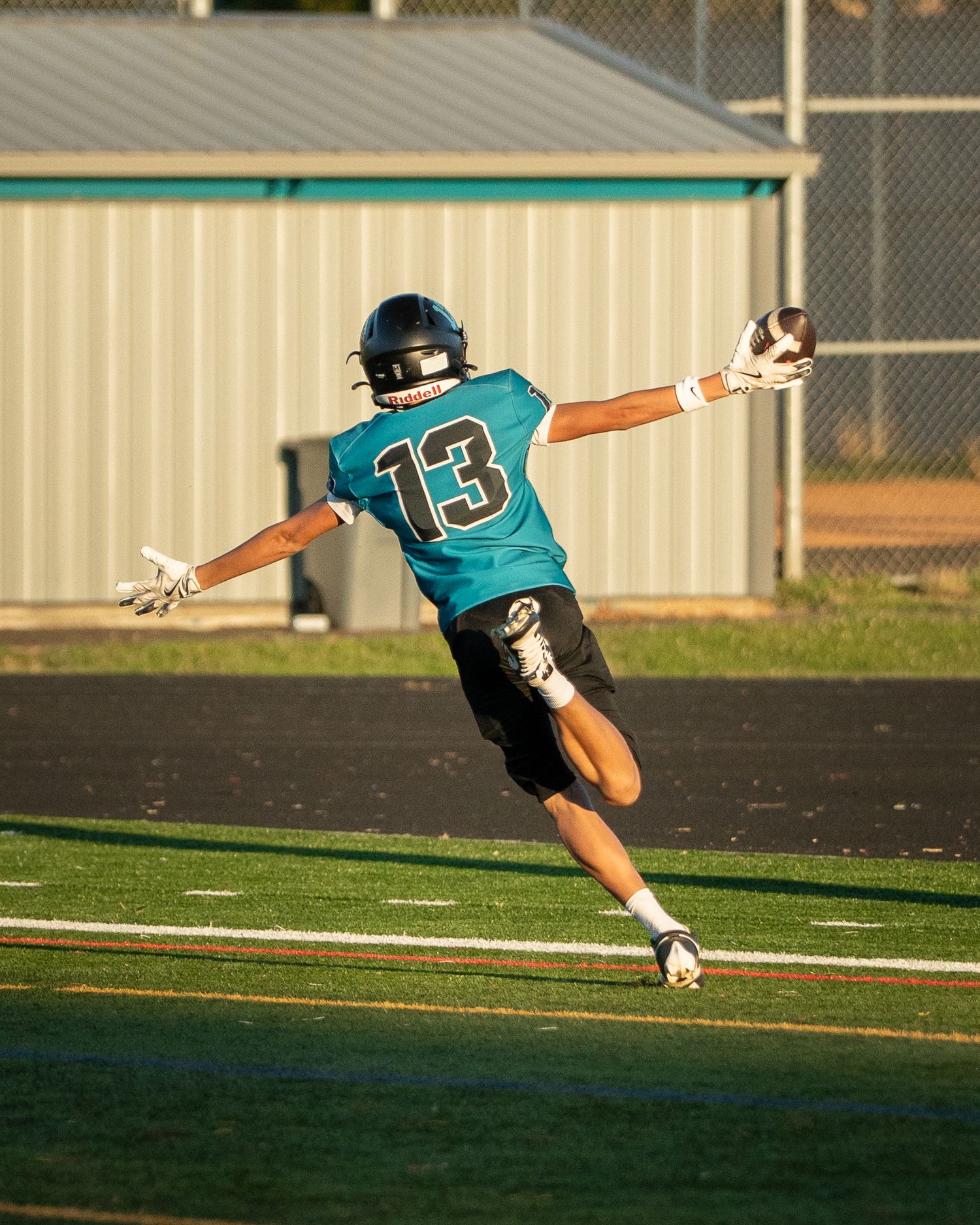 Football player in teal jersey number 13 leaps horizontally across the field during gameplay.