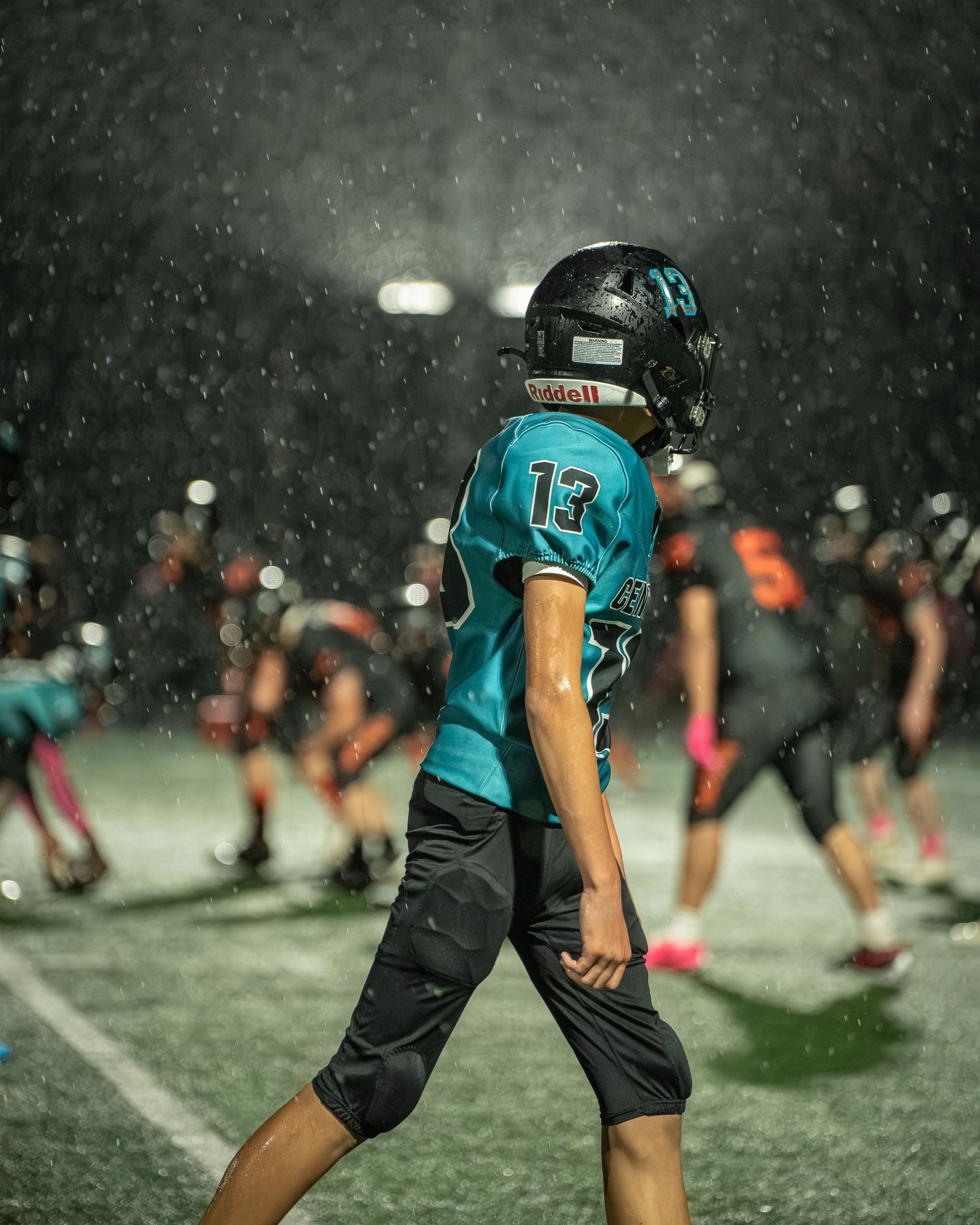 Player in teal jersey stands ready on football field as snow falls during nighttime game.