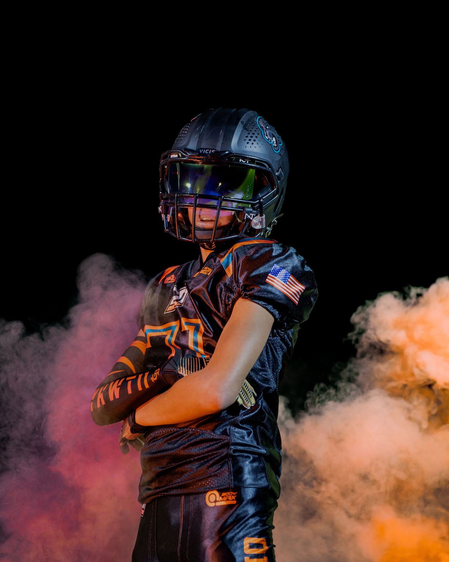 Football player in protective gear poses confidently with crossed arms amid colorful smoke effects on dark background.