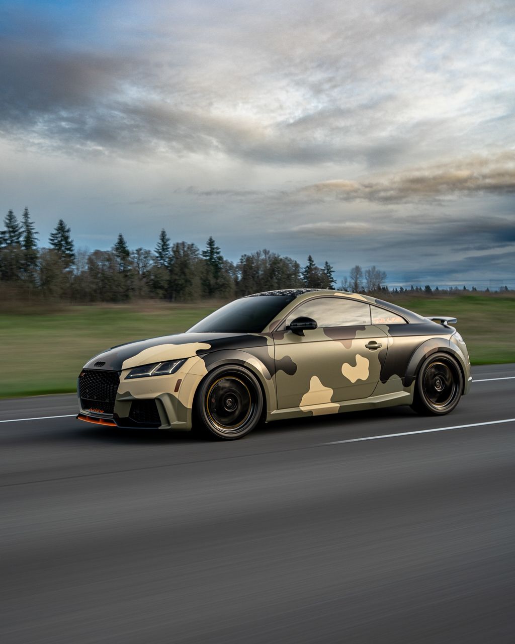 Desert camo wrapped luxury sports car driving on a racetrack under cloudy skies.
