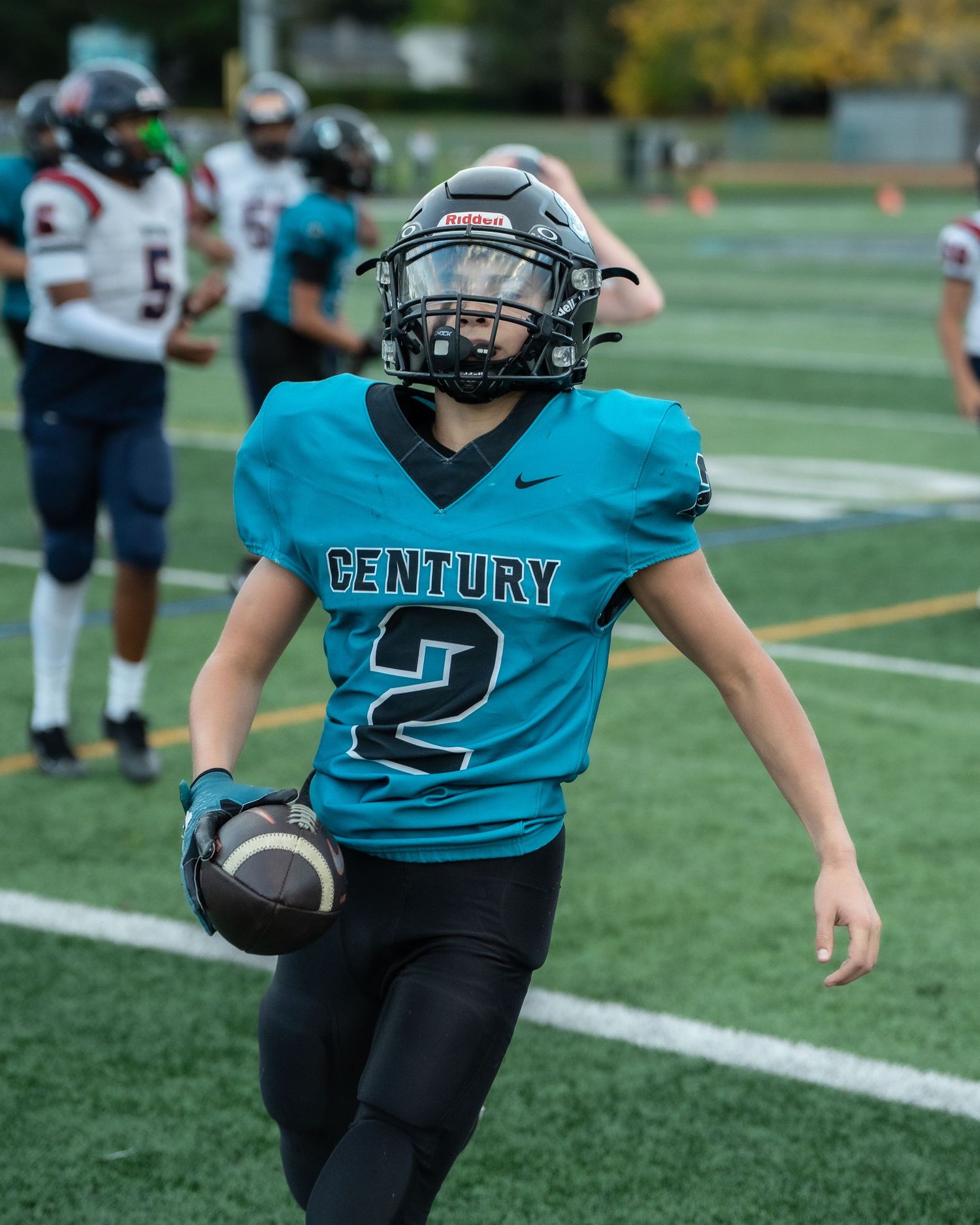 Youth football player in teal Century jersey carrying ball during practice drill.
