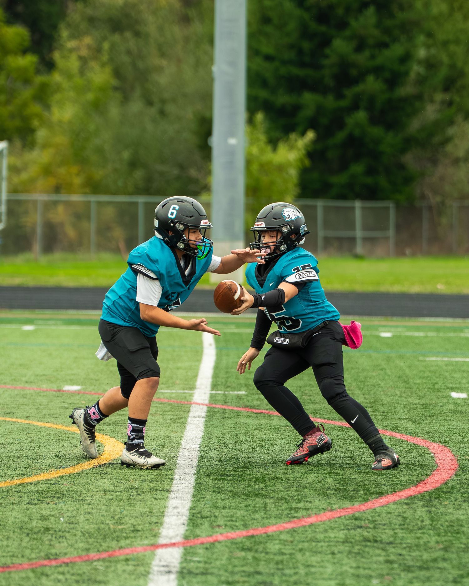 Two players in teal uniforms practice a handoff play during football training.