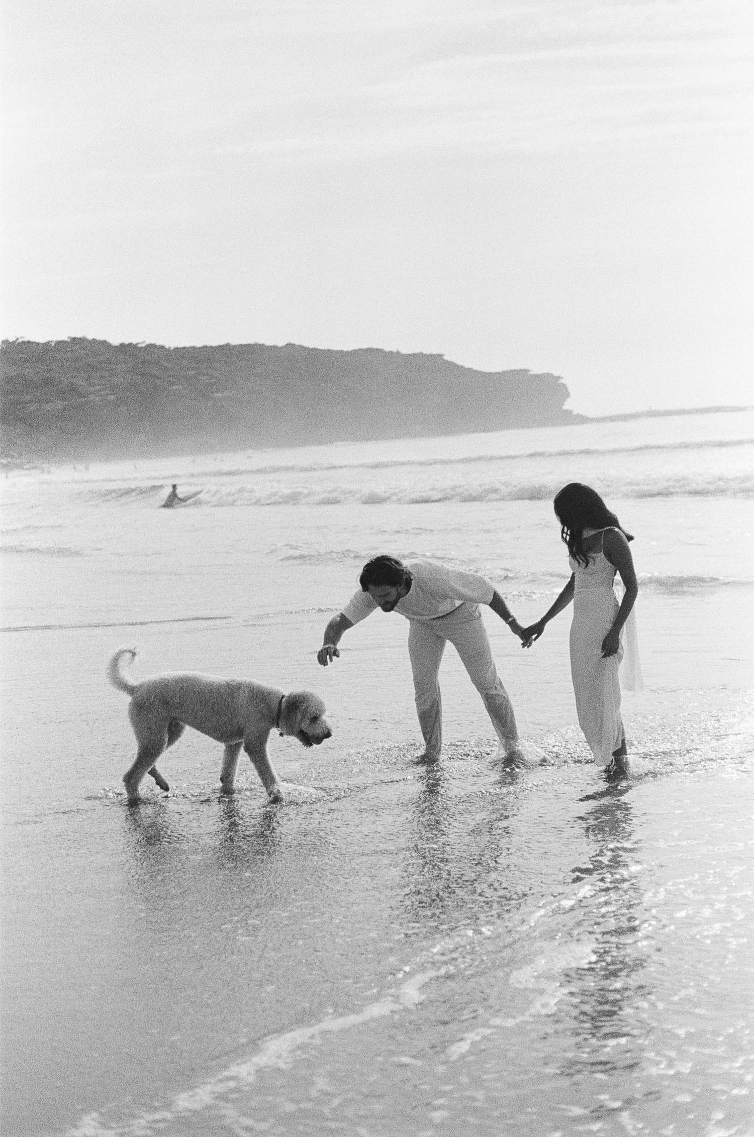 Two people and a dog enjoy a playful moment together at the beach shoreline in black and white photography.