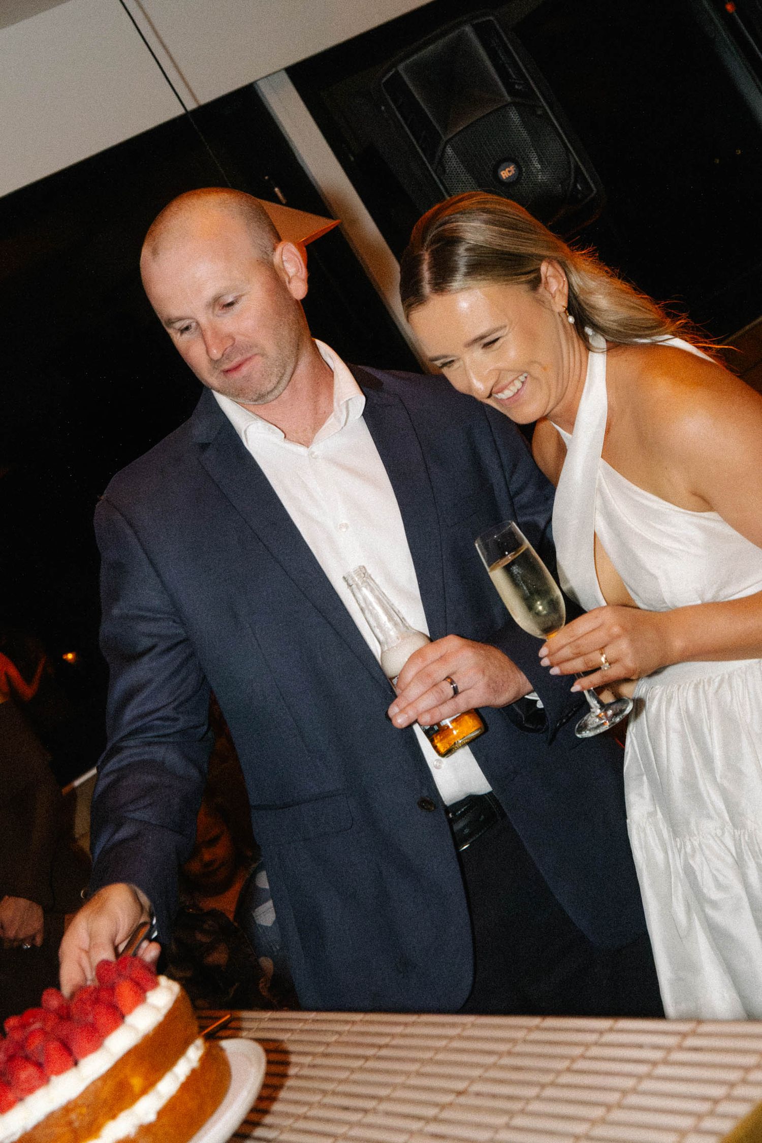A couple in formal wedding attire laughs together while cutting their wedding cake at the reception.