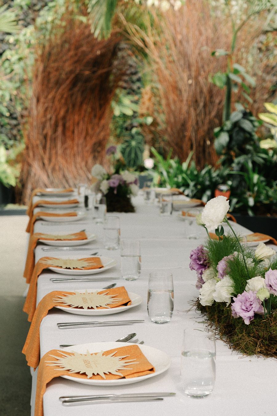 Elegant table setting with orange napkins and place settings arranged under dried pampas grass decor at outdoor event.