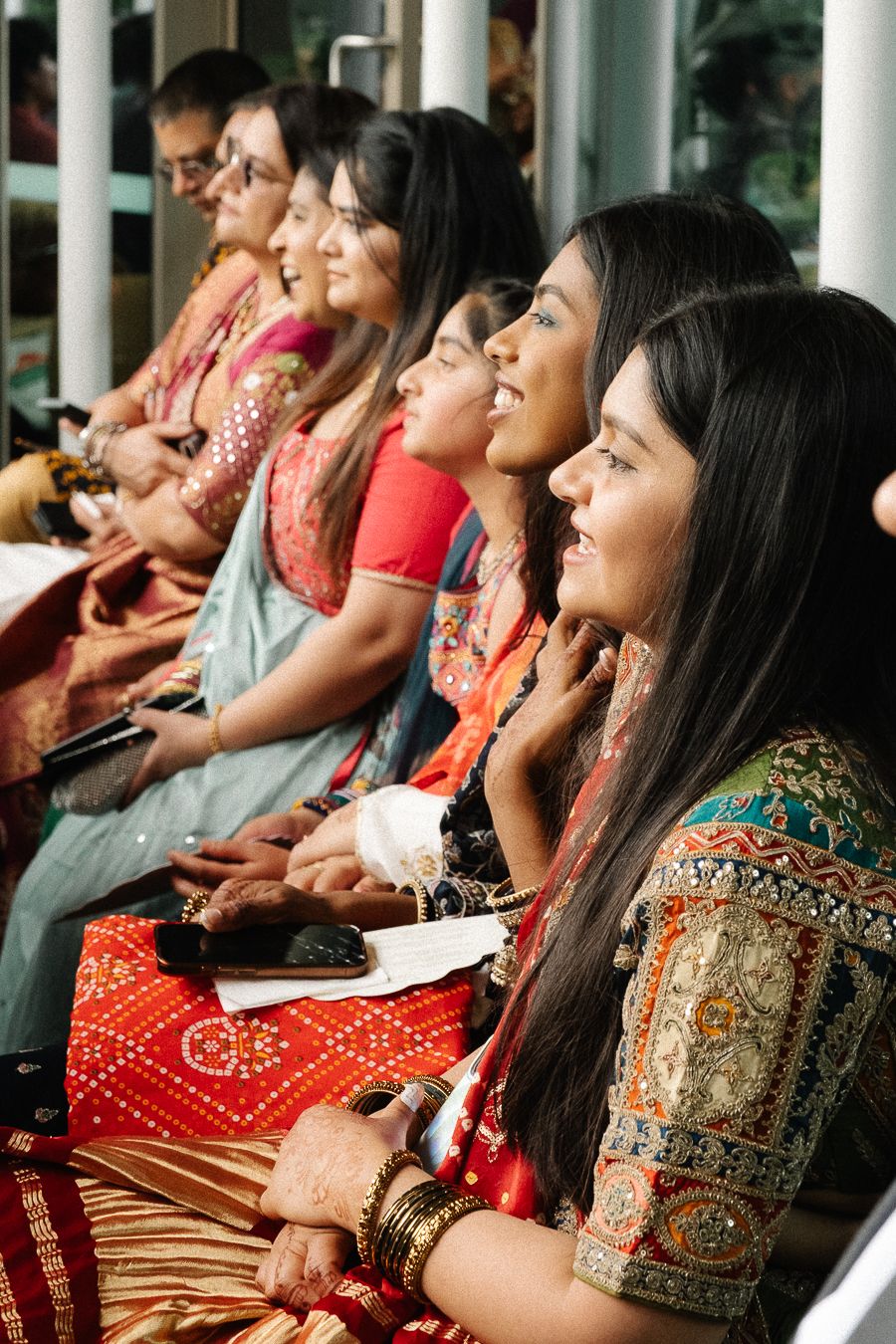 A group of women in traditional Indian attire sit together and smile while wearing colorful saris and jewelry.