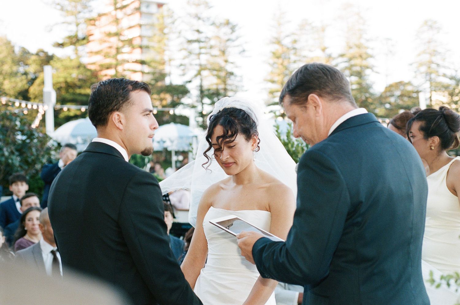 A bride in a white gown and veil exchanges vows with a groom in a dark suit as an officiant reads during an outdoor wedding ceremony.