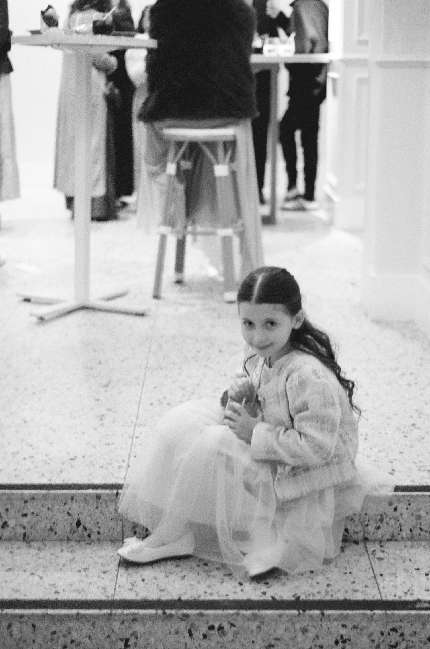 Smiling young girl in a tulle skirt and cardigan sits on a terrazzo step, holding a small item, with guests gathered at a table behind her.