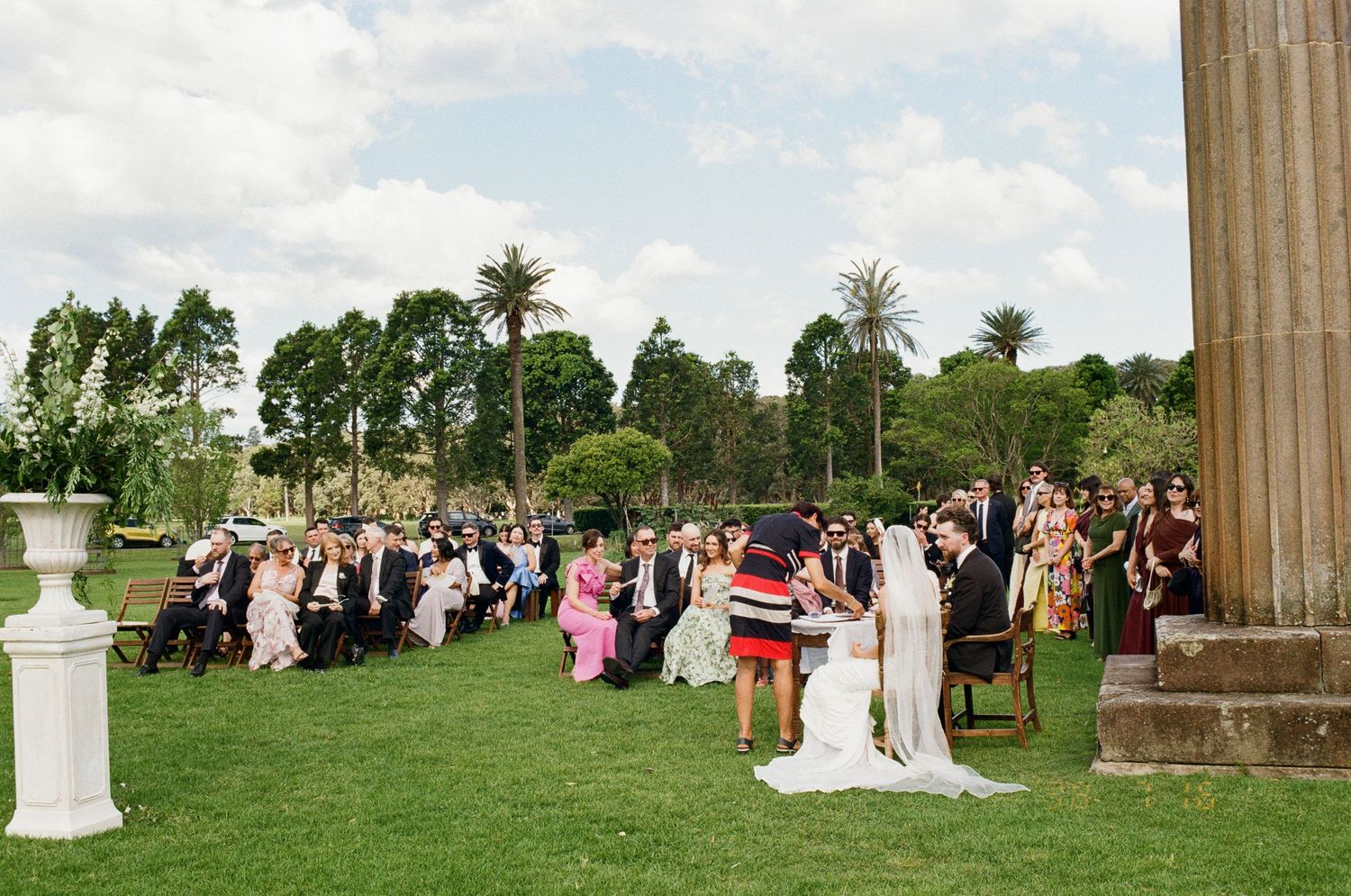Wedding guests seated on chairs watch a bride in a white gown and groom exchange vows on a sunny green lawn beside a stone column.