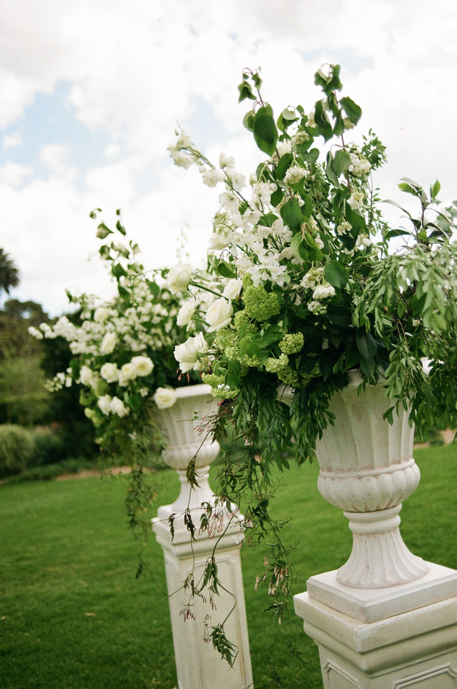 White roses and green foliage overflow from classic stone urns on pedestals, creating an elegant garden wedding ceremony display.