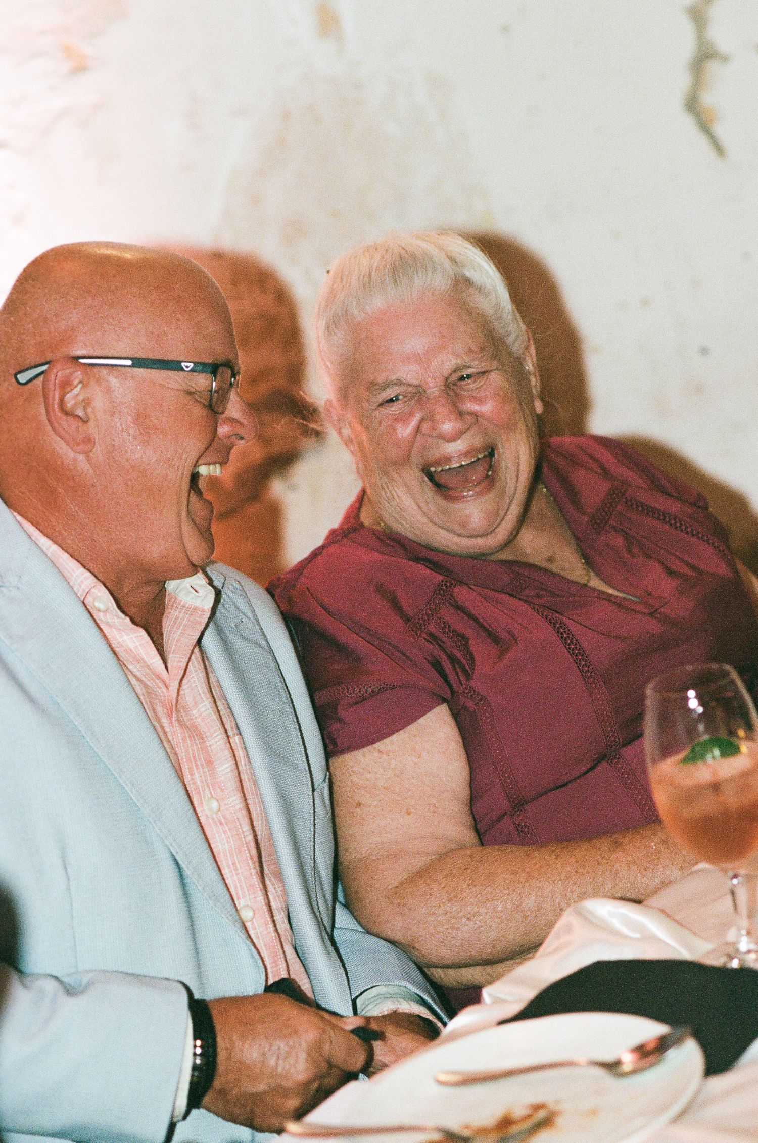 Two older adults laugh heartily together at a dinner table, one in a light blue blazer and glasses, the other in a maroon top, with a cockta