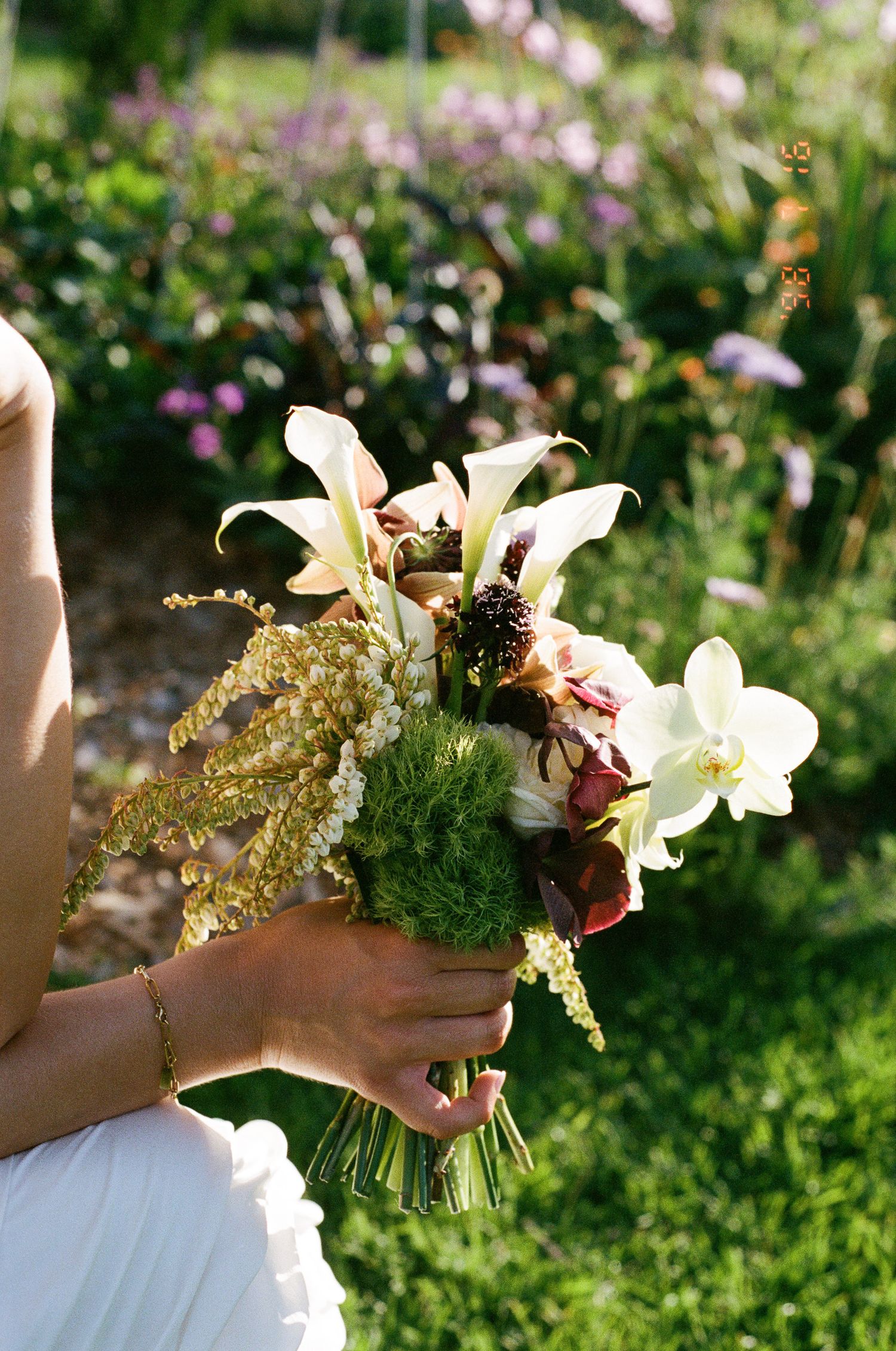 Bride holding a wildflower bouquet of white orchids, calla lilies, green moss and golden astilbe in a sunny garden.