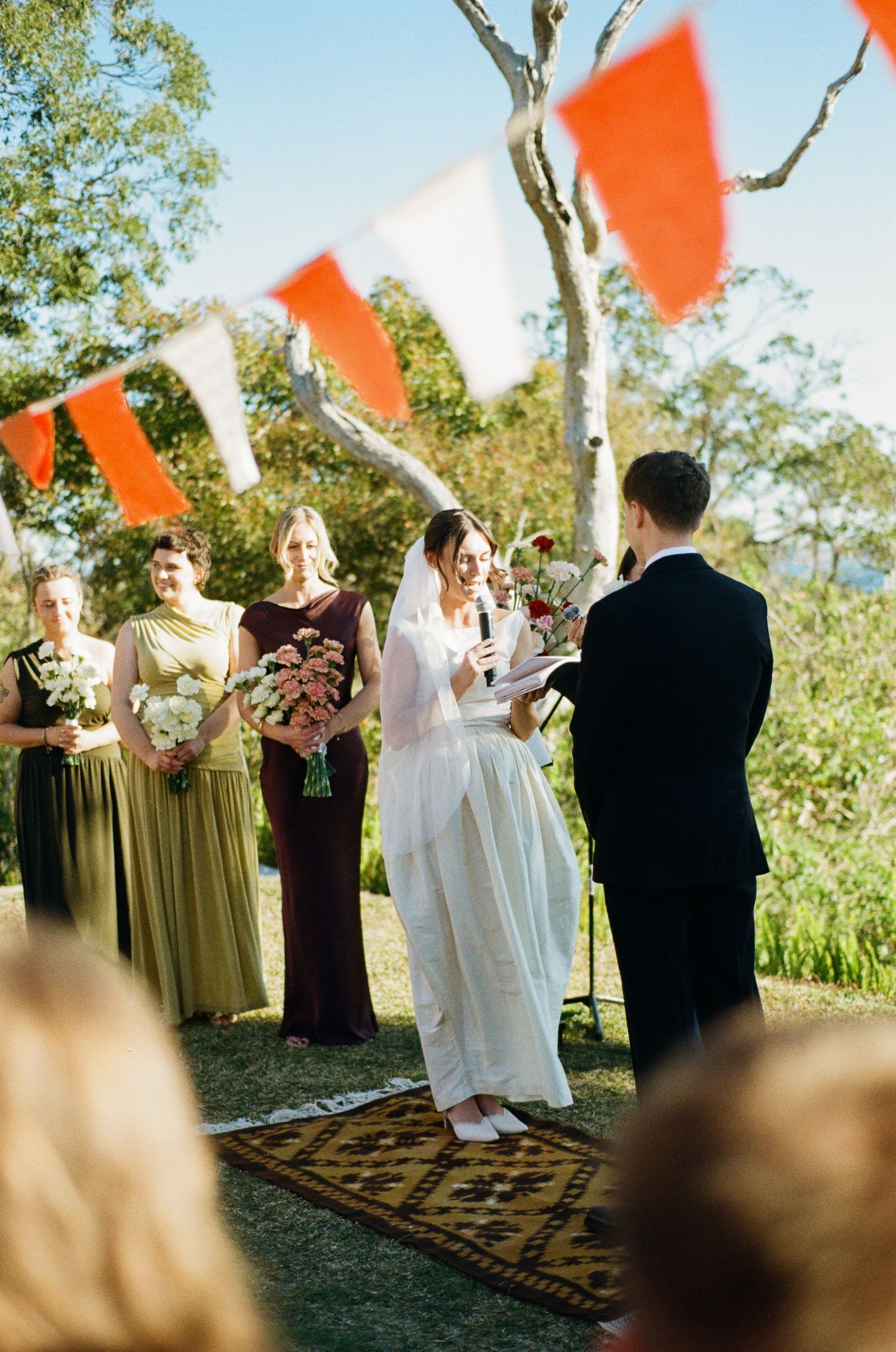 Bride in a white gown speaks her vows into a microphone as bridesmaids with bouquets and colorful bunting look on at an outdoor wedding.