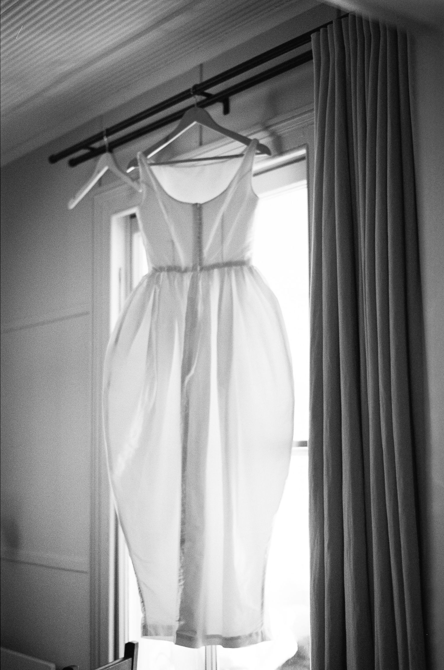 Black and white photo of a white wedding dress with a fitted bodice hanging from a window rod beside a draped curtain in soft natural light.