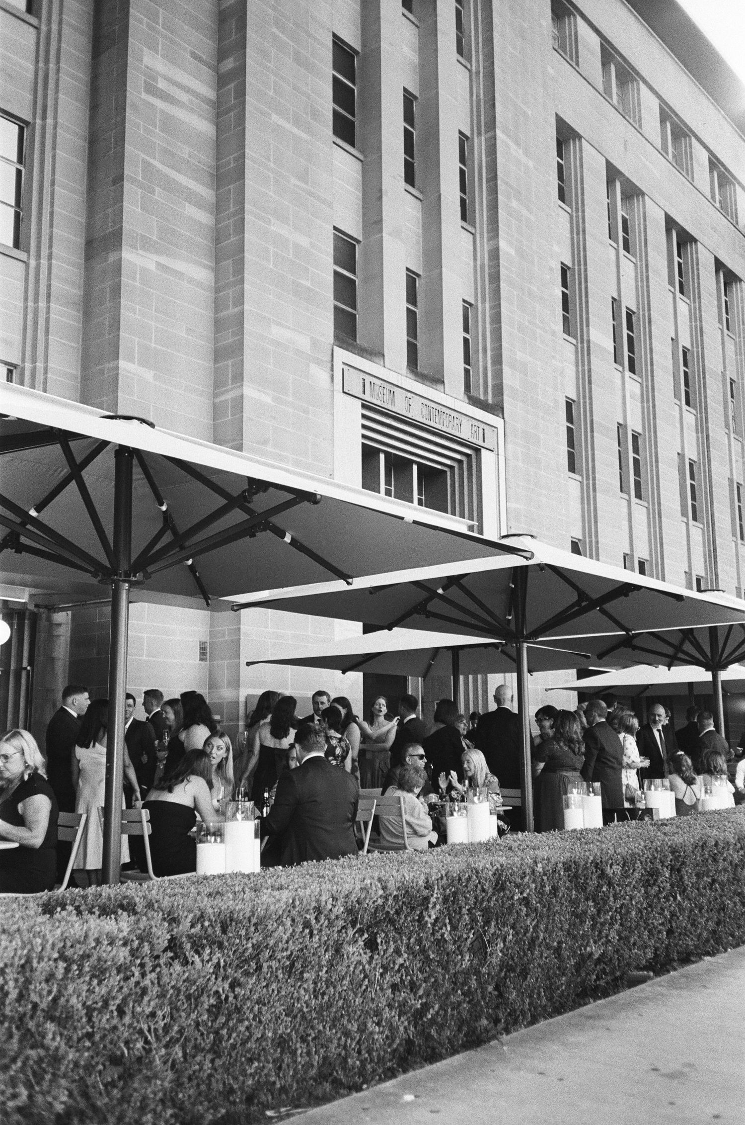 Black and white photo of guests gathering at outdoor tables beneath large patio umbrellas beside a trimmed hedge and a tall stone building.