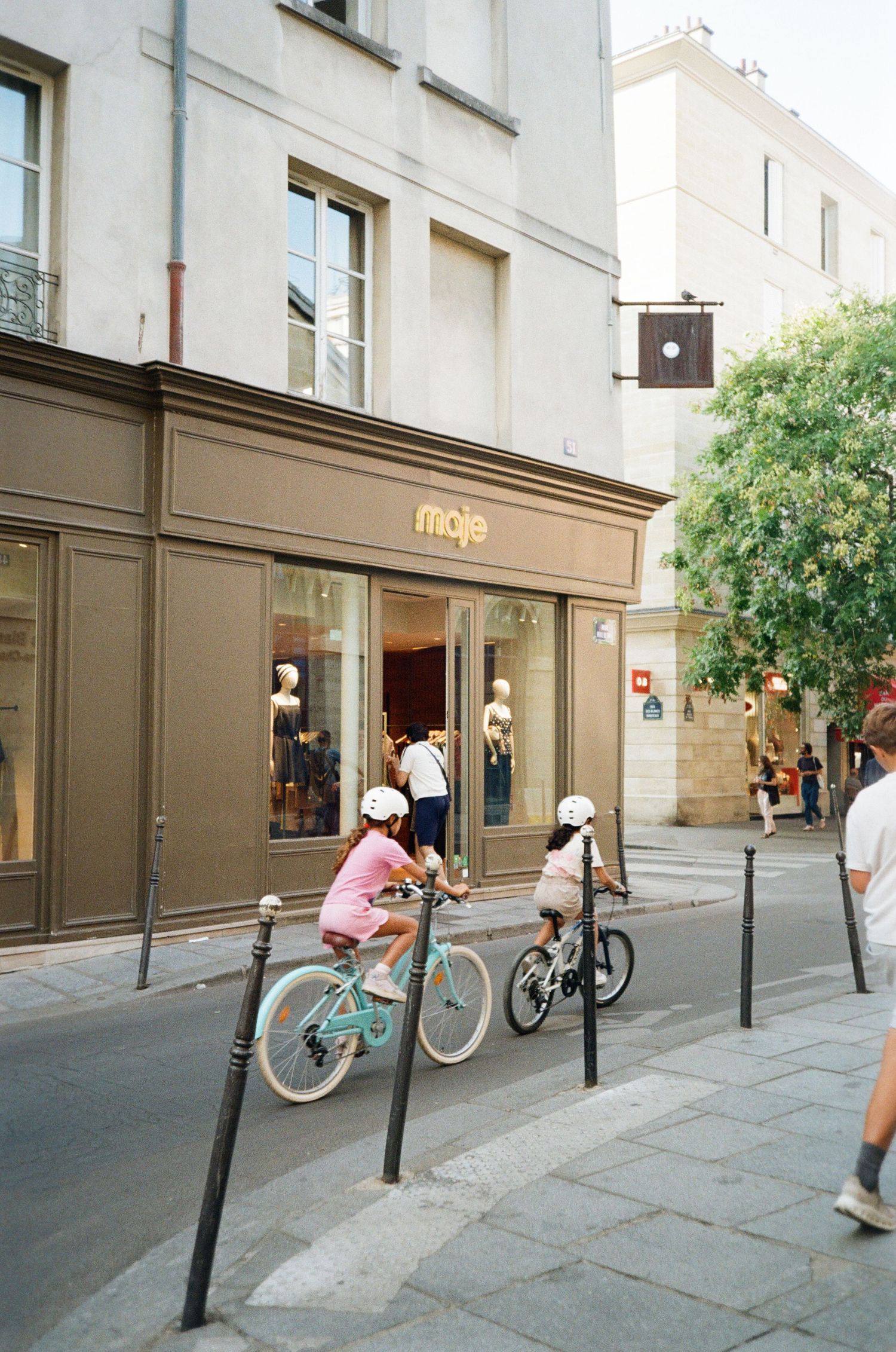 Two children in helmets ride bicycles past a Maje storefront with mannequins in the window on a sunny Parisian street.