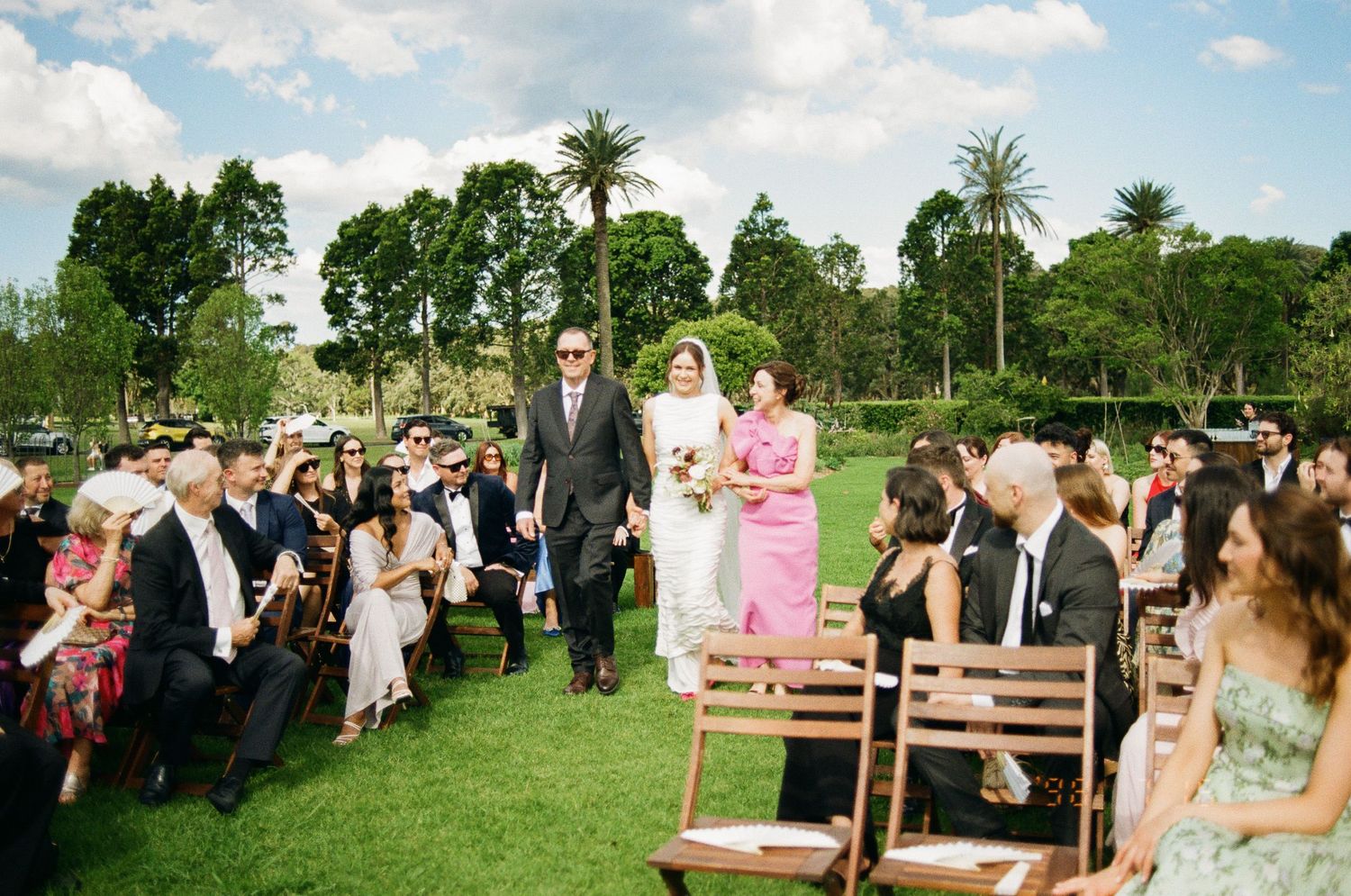 Bride in white gown walks arm in arm with a man in a gray suit down the grass aisle of an outdoor wedding as seated guests watch.