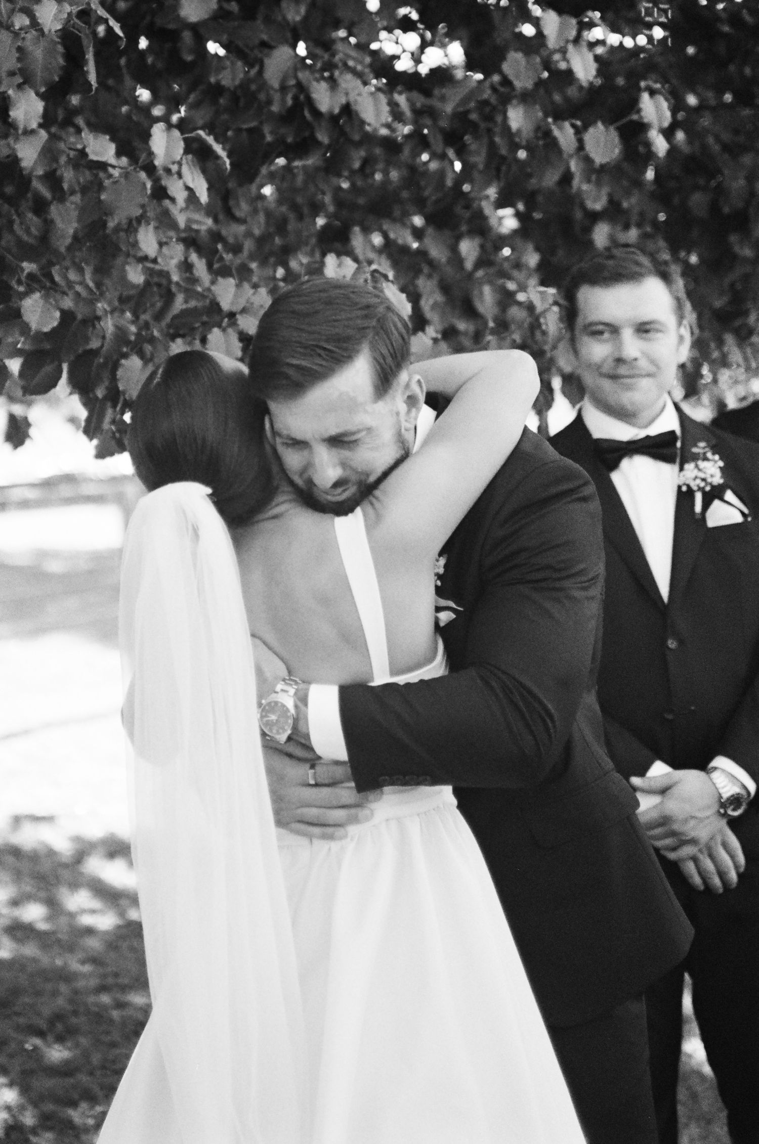 Emotional groom in a black tuxedo embraces his bride in a white gown and veil during an outdoor wedding ceremony as a groomsman smiles.