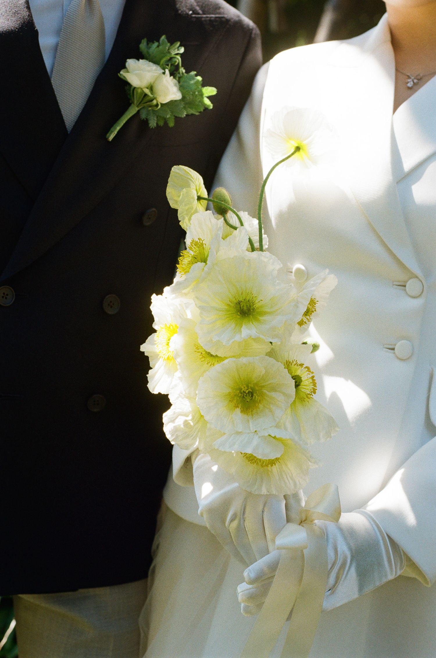 Bride in a white jacket and gloves holds a bouquet of cream poppies beside a groom in a dark suit with a white boutonniere.