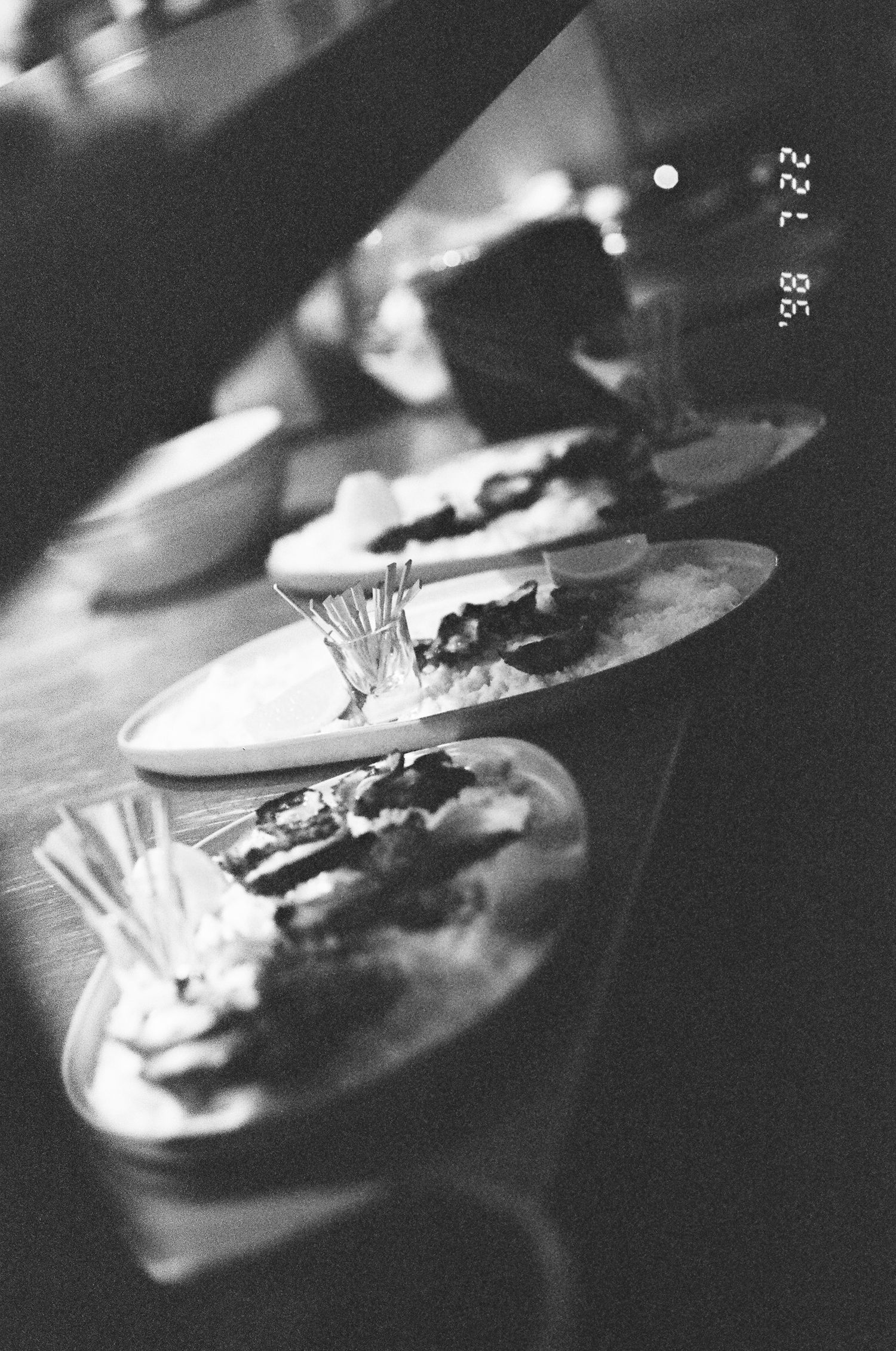 Black and white photo of three oval plates of food with toothpick bundles, arranged in a row on a dark bar counter.