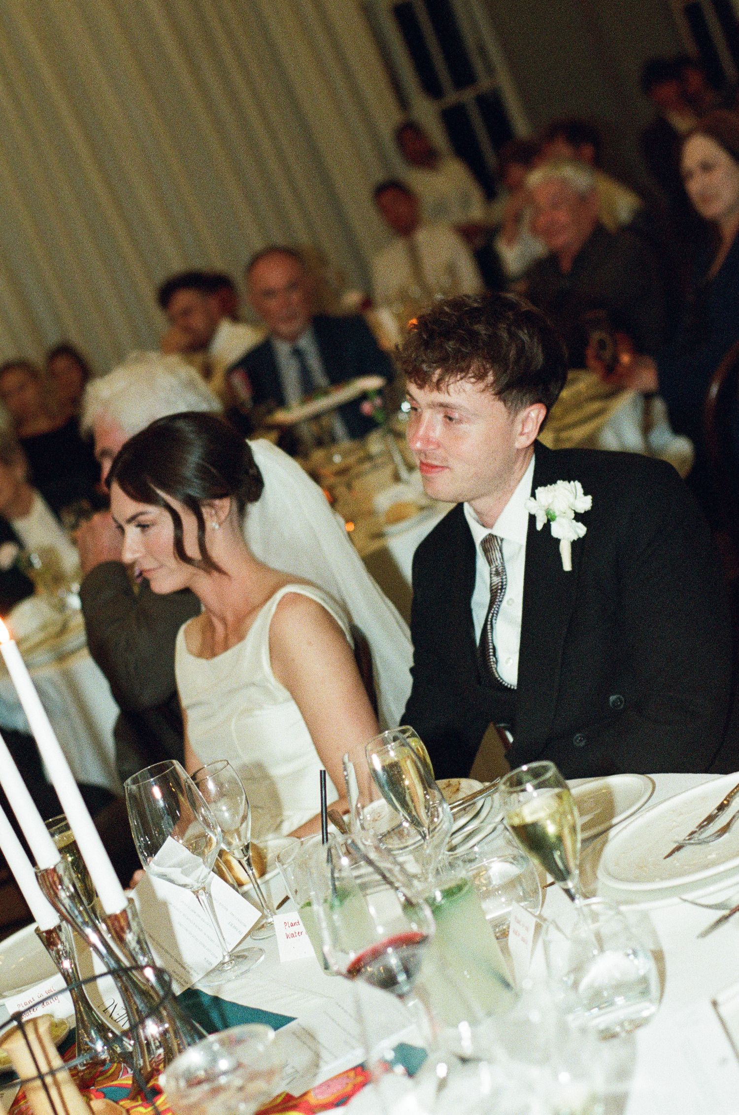 Bride in a white dress and groom in a black suit with a boutonniere sit at a candlelit wedding reception table as guests look on.