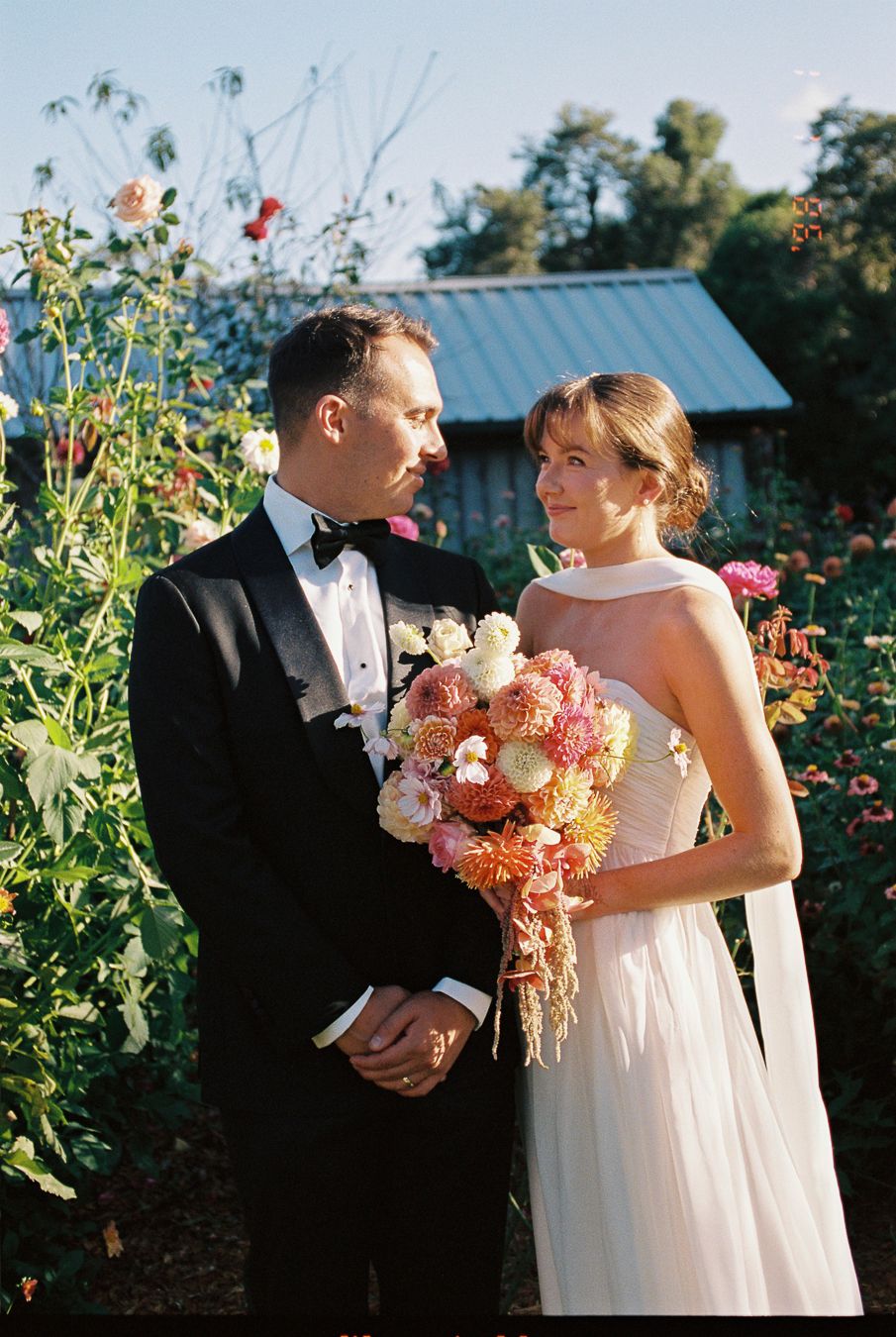 Bride holding colorful bouquet of pink and orange flowers stands with groom in black tuxedo in garden setting.