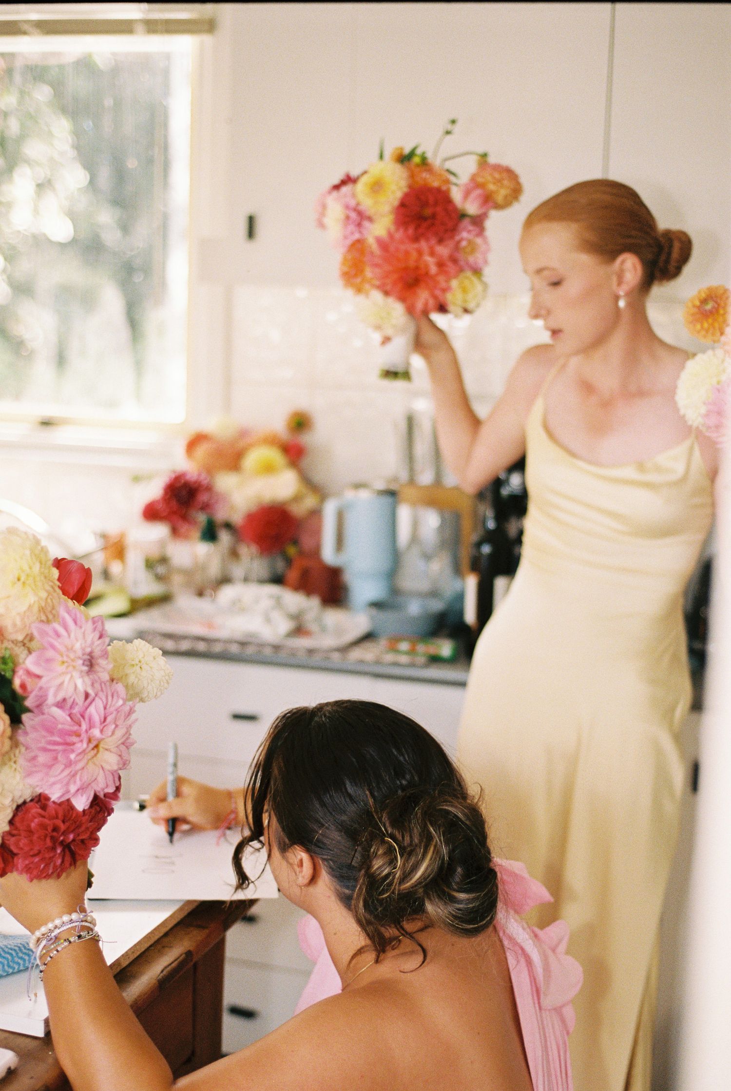 Bridesmaid in a yellow satin dress holds a dahlia bouquet in a bright kitchen while another woman in pink writes at a table with flowers.
