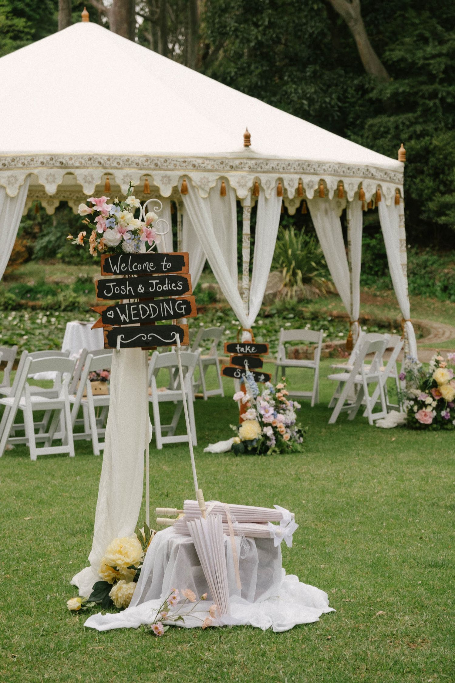 Elegant outdoor wedding setup with white tents, floral arrangements, and chalkboard welcome signs on a lush green lawn.