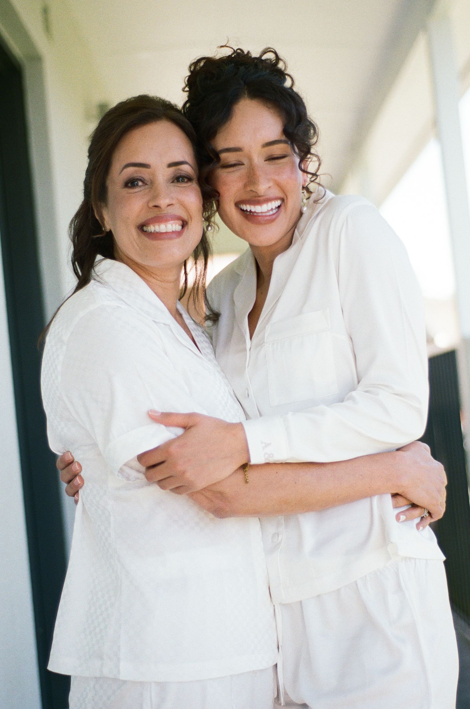 Two smiling women in matching white button-up pajama sets embrace warmly on a sunlit balcony, sharing a joyful moment together.