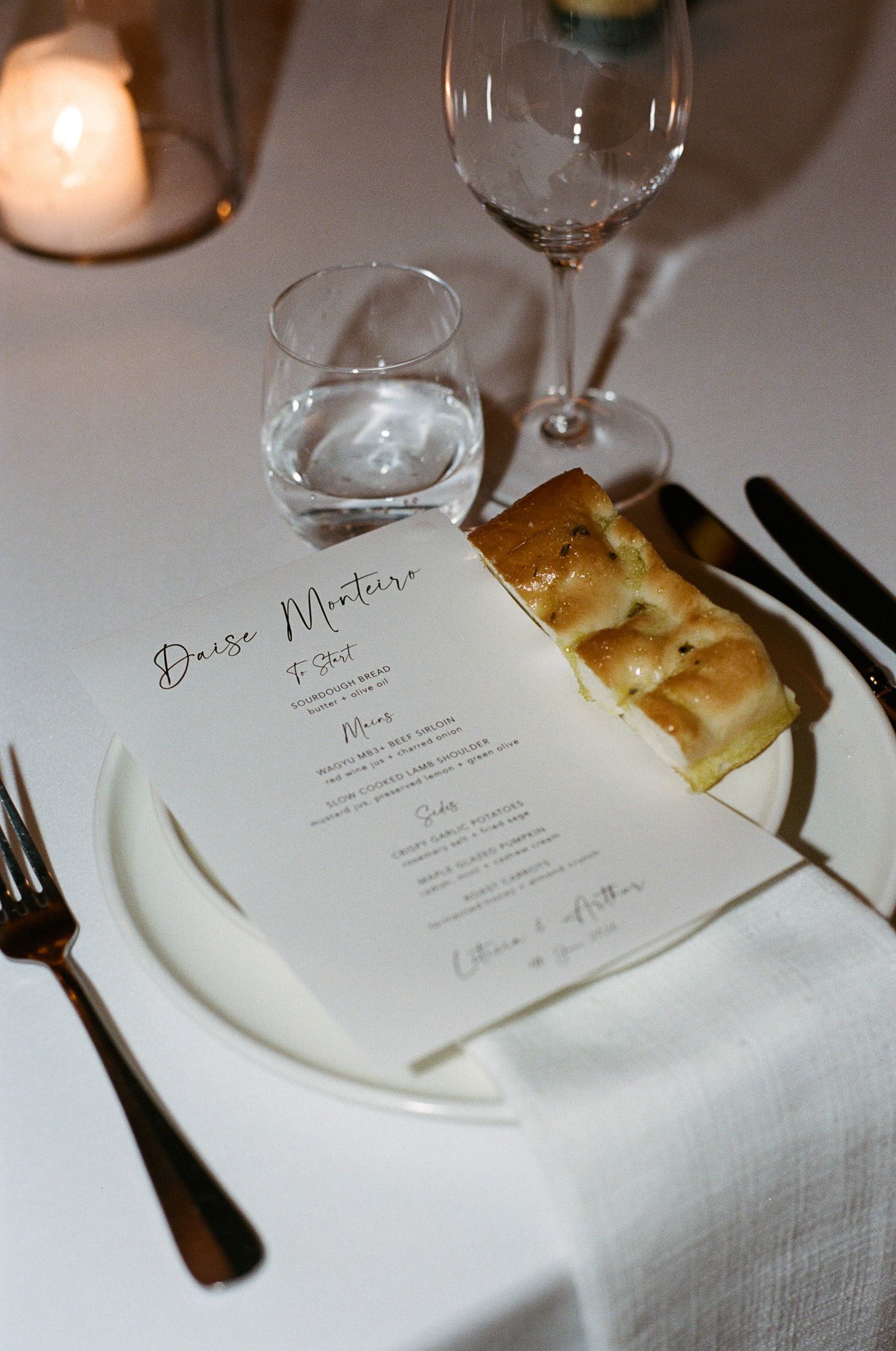Wedding reception place setting with a printed menu card, focaccia slice, white napkin, wine glass, water glass, and candlelight.