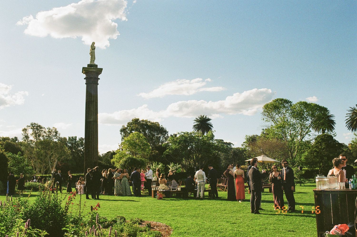 Guests in formal attire gather on a sunny green lawn near a tall stone column topped with a statue, surrounded by trees under a blue sky.