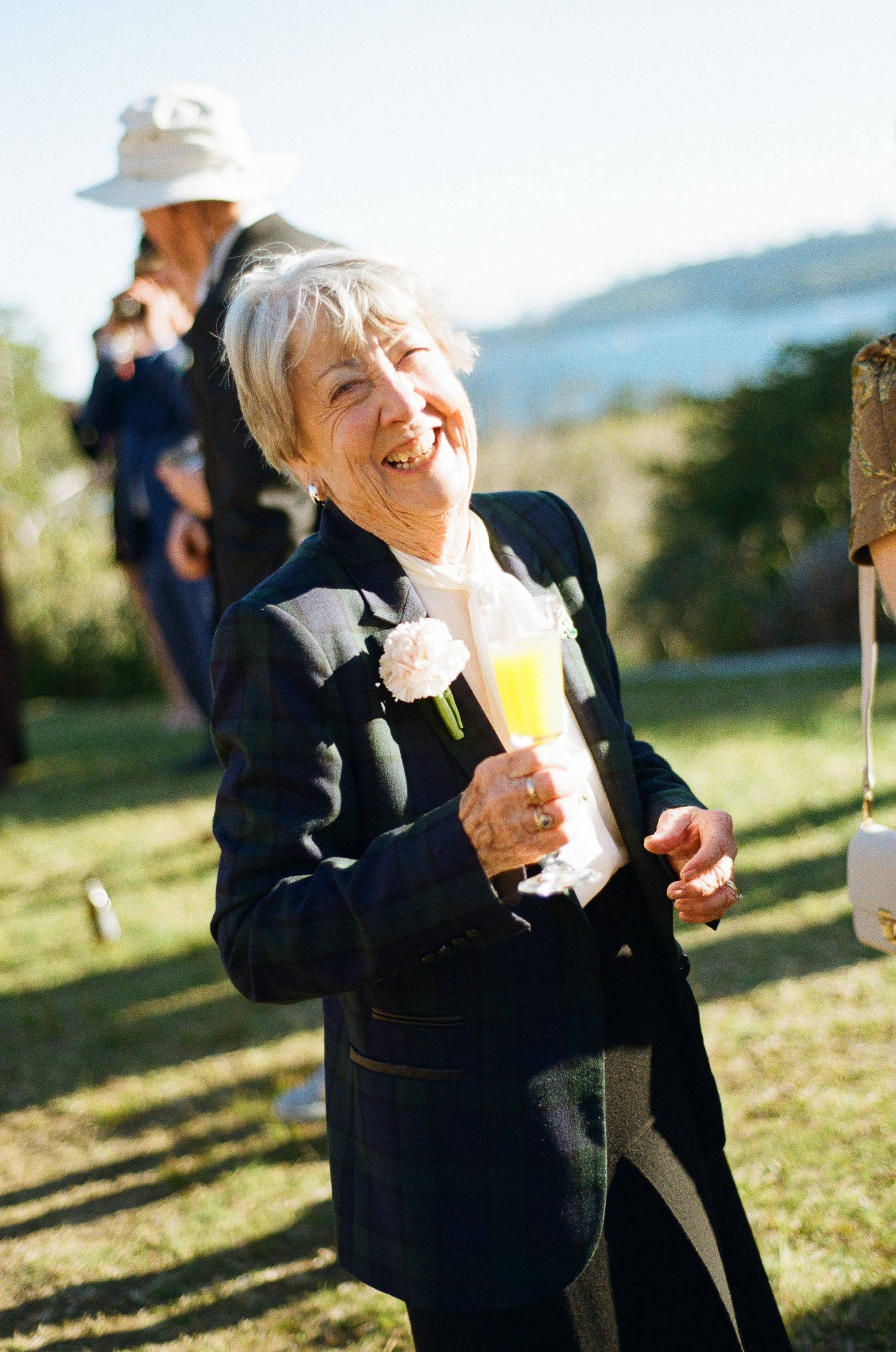 Smiling older woman with short gray hair holds a champagne flute at an outdoor garden party, wearing a plaid blazer with a pink boutonniere.