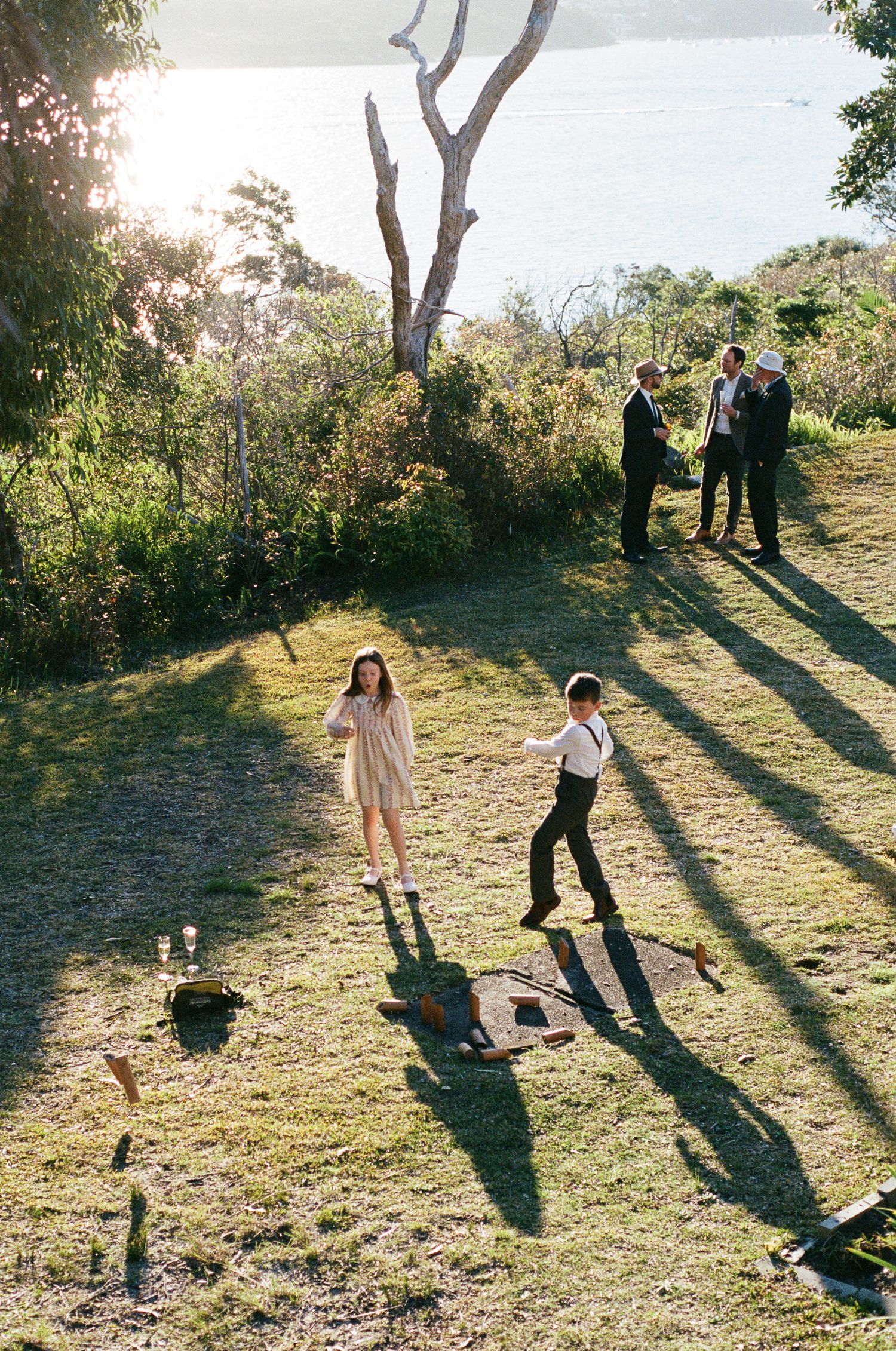 Two children play a lawn game on a sunlit grassy hillside while three men in formal attire chat nearby, with a sparkling bay behind them.