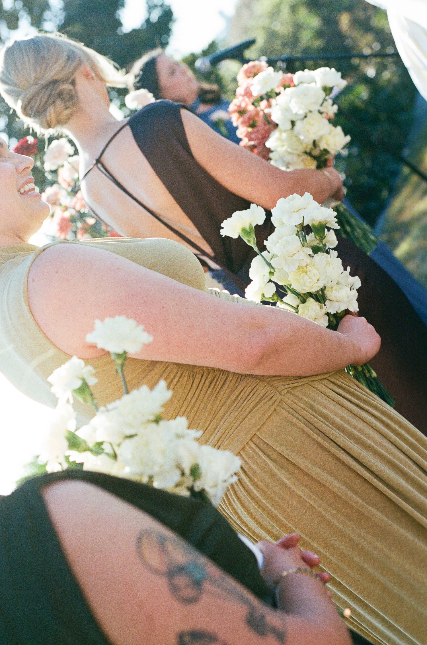 Bridesmaids in gold and black dresses hold white flower bouquets and laugh together outdoors at a sunlit wedding ceremony.