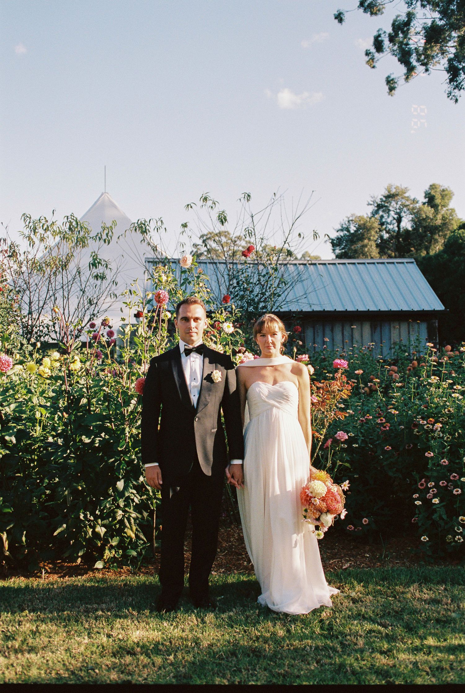A bride in a flowing white gown holding a bouquet stands beside a groom in a black tuxedo in a blooming flower garden at an outdoor wedding.