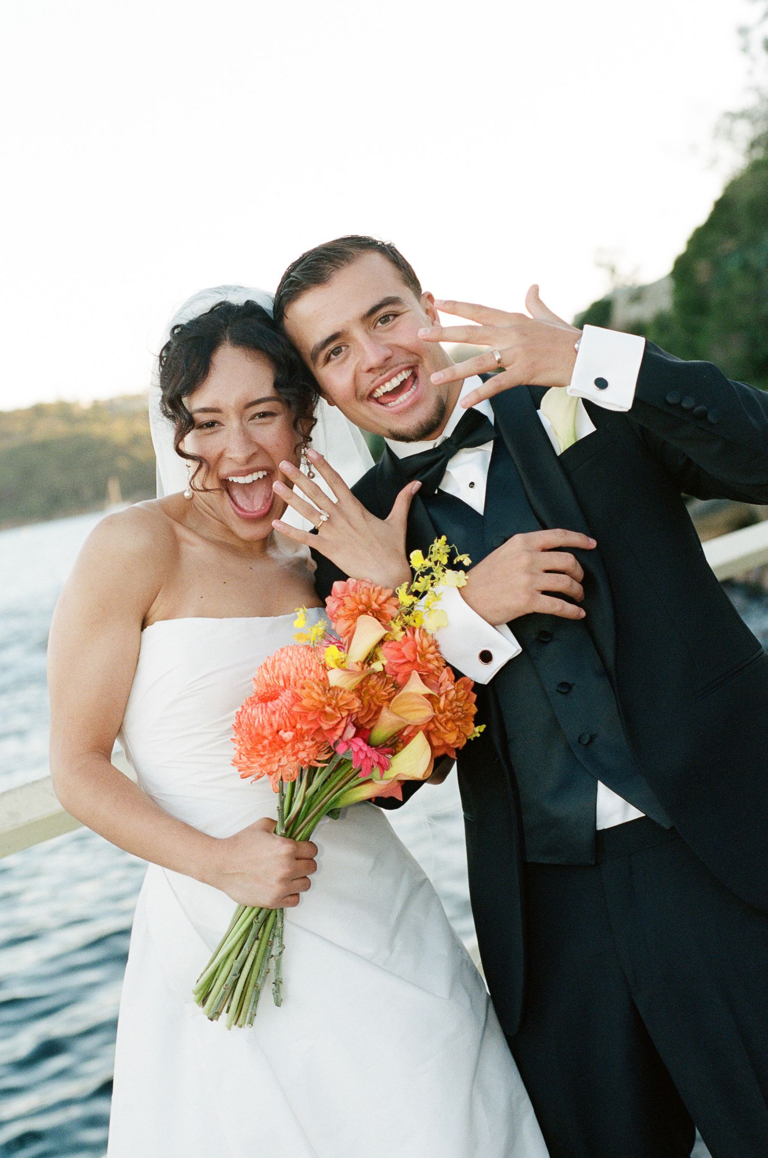 Joyful bride and groom laughing and showing off their wedding rings by the water, with an orange and pink bridal bouquet.